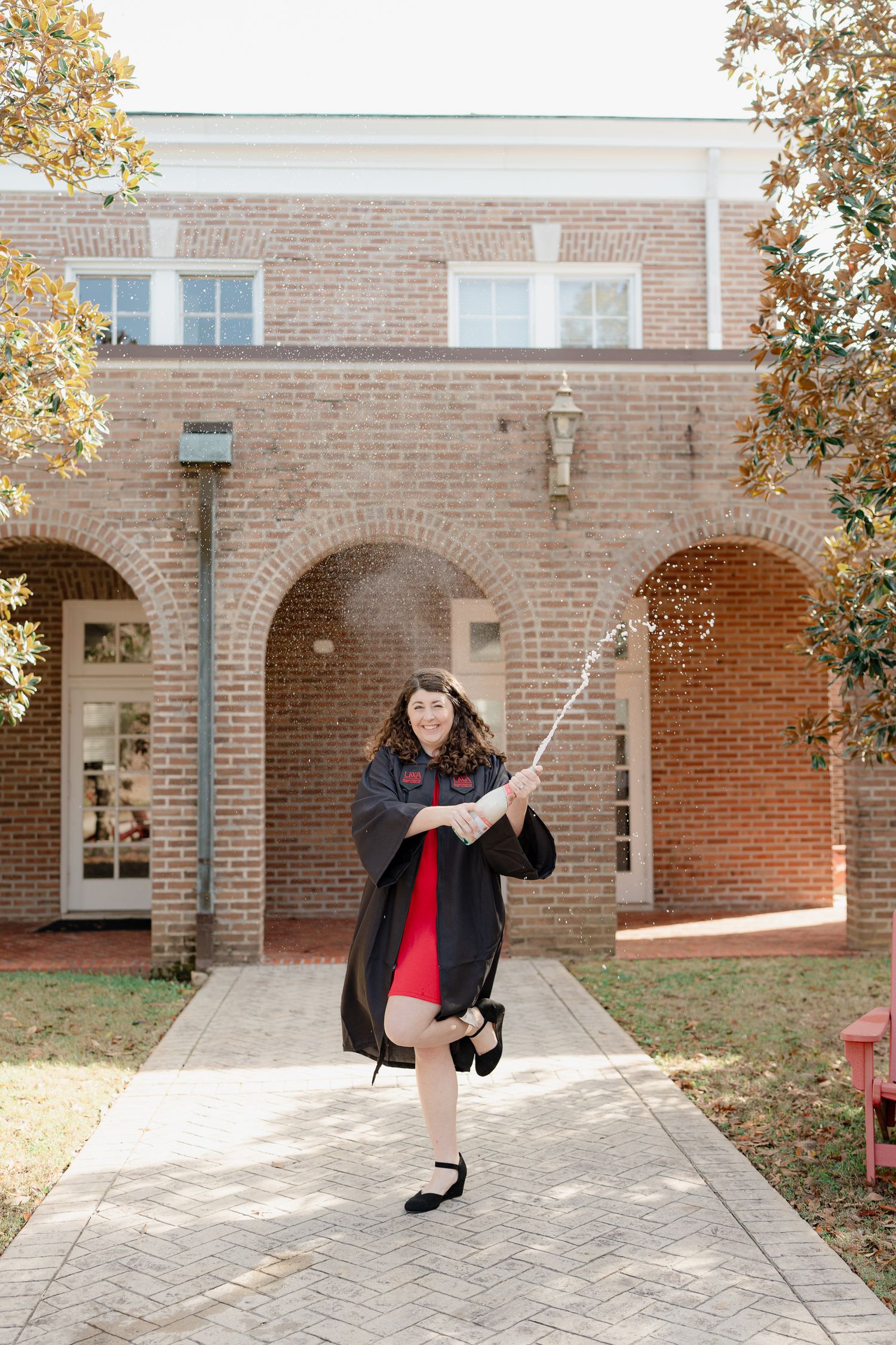 Graduate in black cap and gown poses playfully with selfie stick in front of brick archway on college campus.