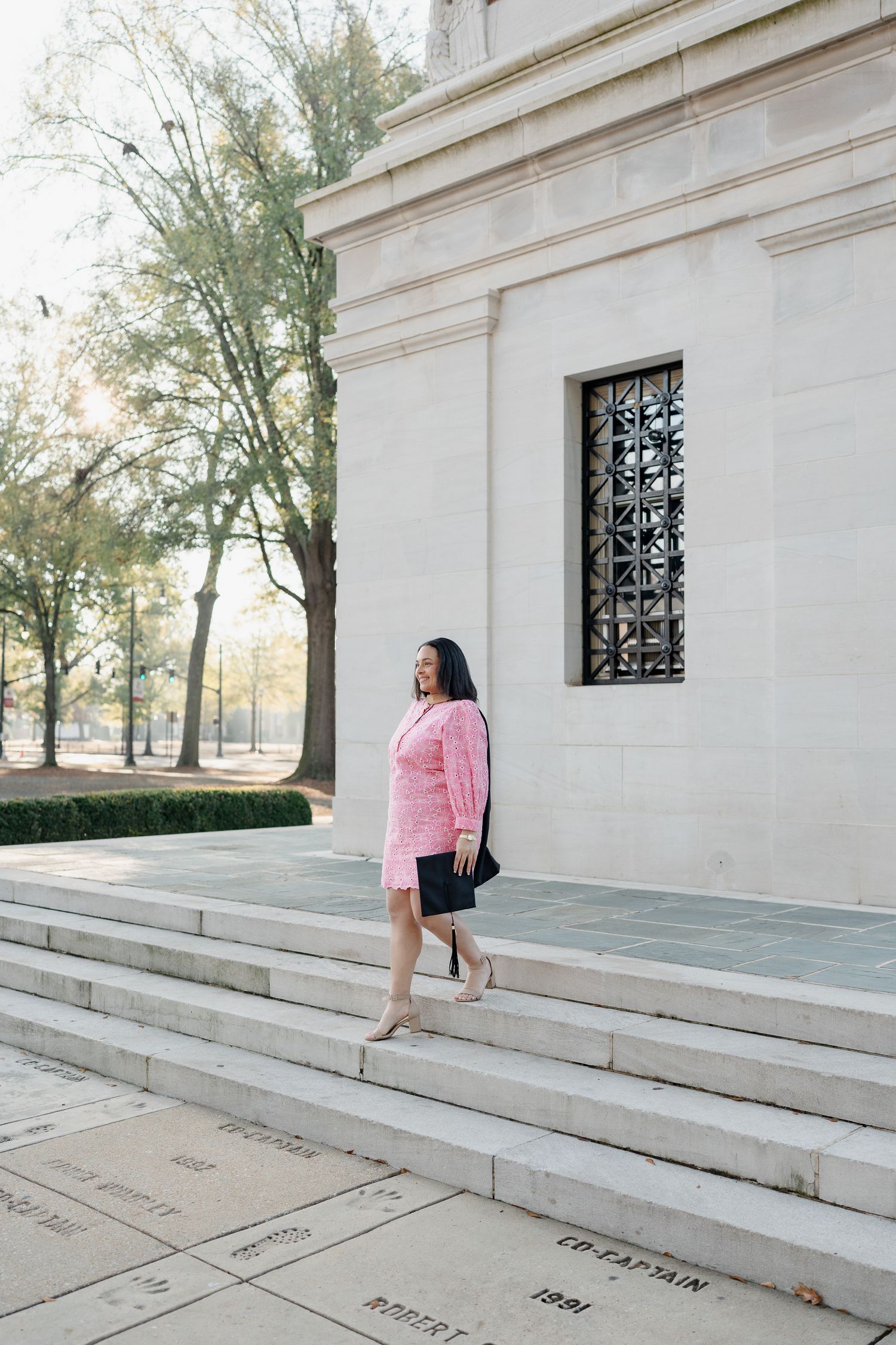 Graduate in pink sweater poses on white stone steps of classical campus building.