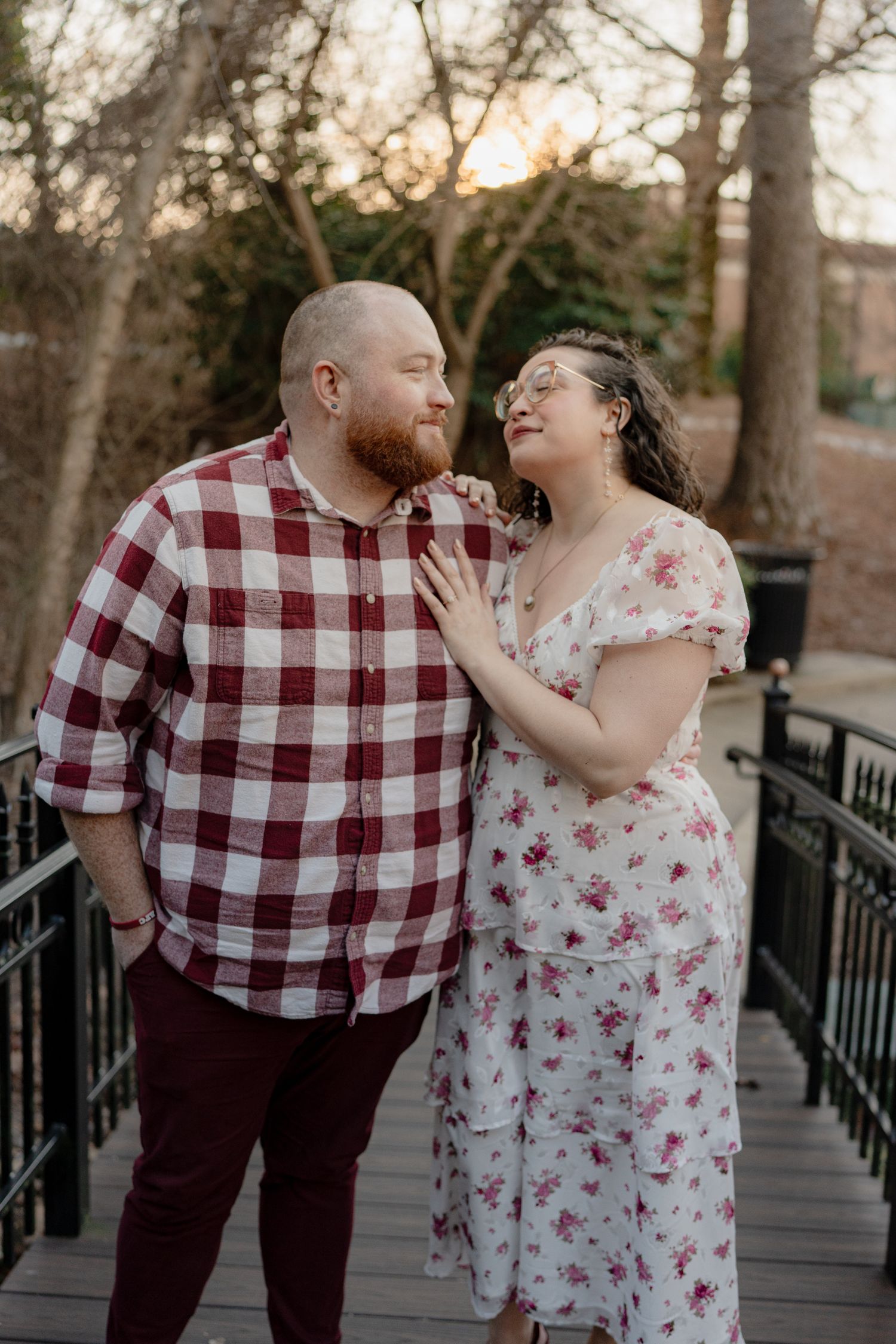 A couple shares a tender moment on a wooden bridge at sunset, with her wearing a floral dress and him in a plaid shirt.