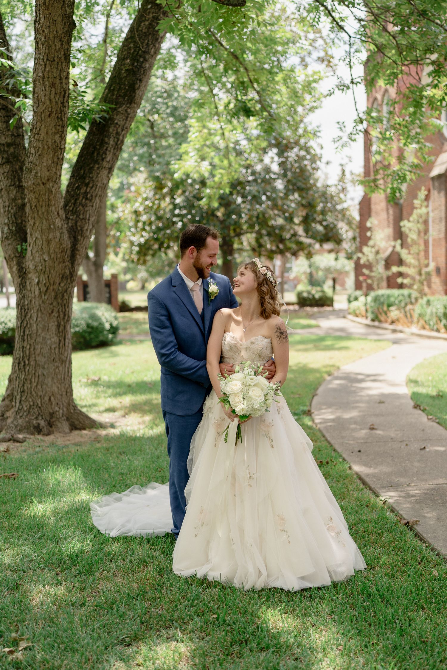 A romantic wedding couple poses under towering trees along a stone pathway in an outdoor garden setting.