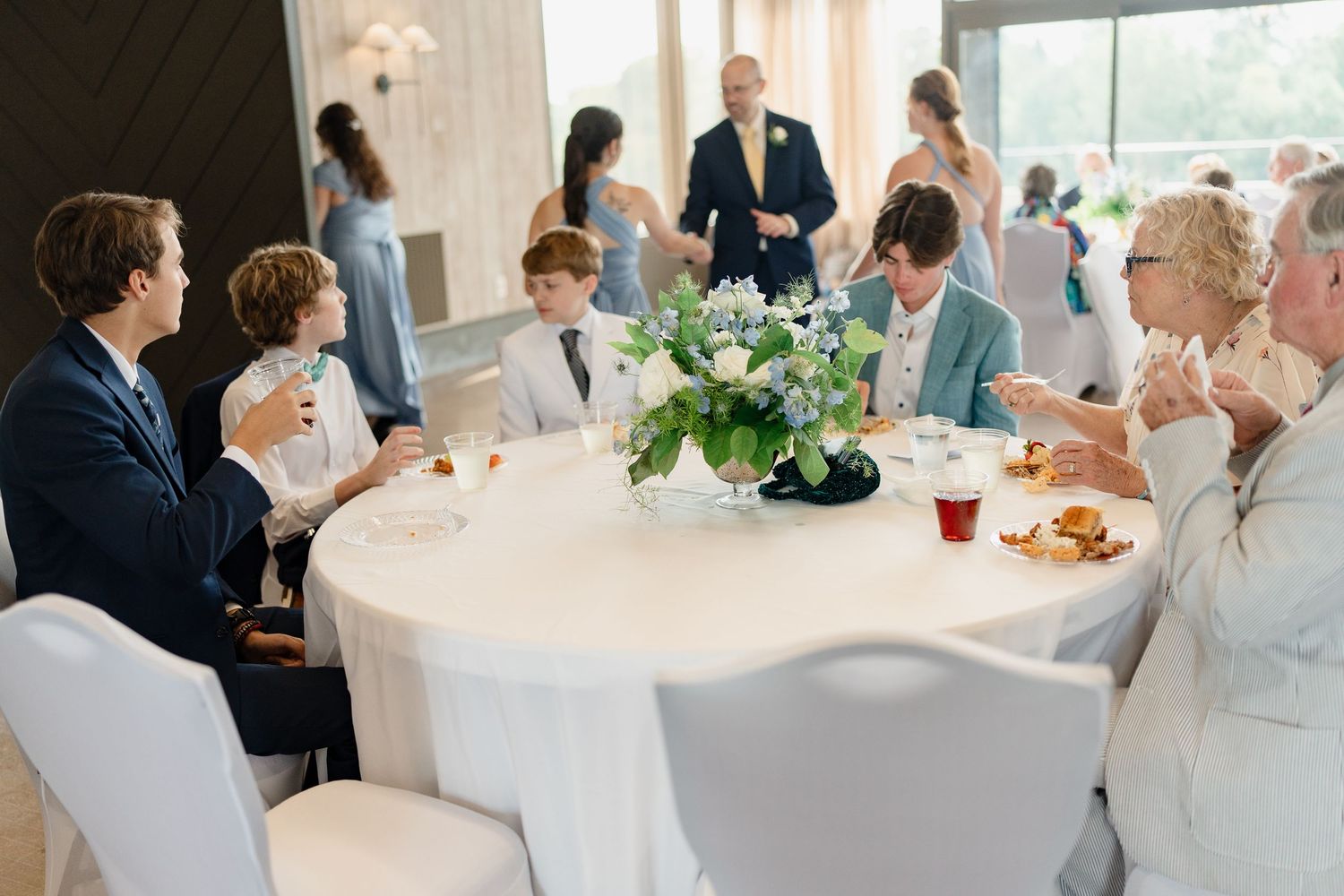 Wedding guests gather at elegant round tables draped in white linens during an indoor reception celebration.