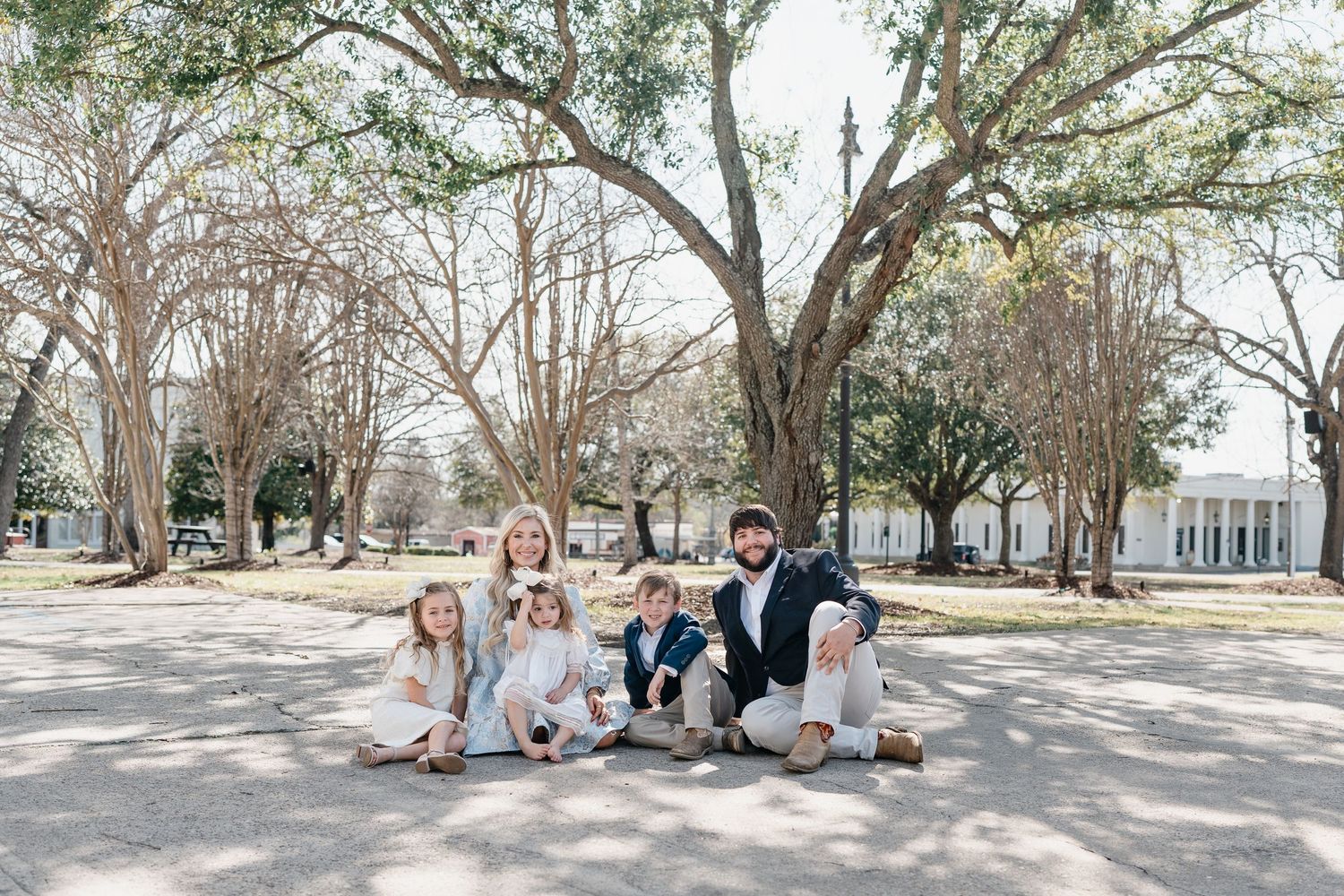 Family group sitting together on ground under large oak trees in coordinated outfits.