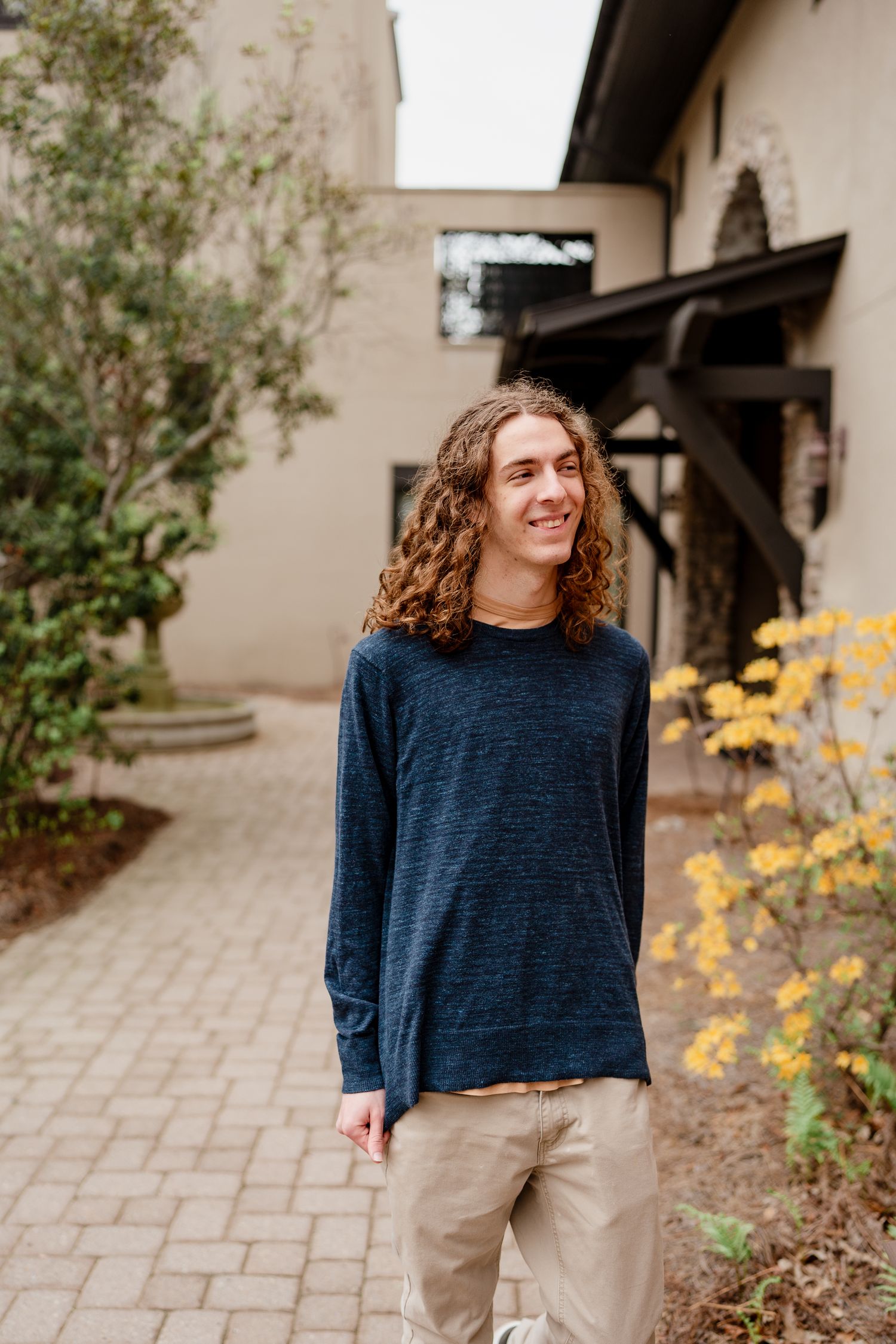 Person in navy sweater and khaki pants standing on brick path with yellow flowers nearby.