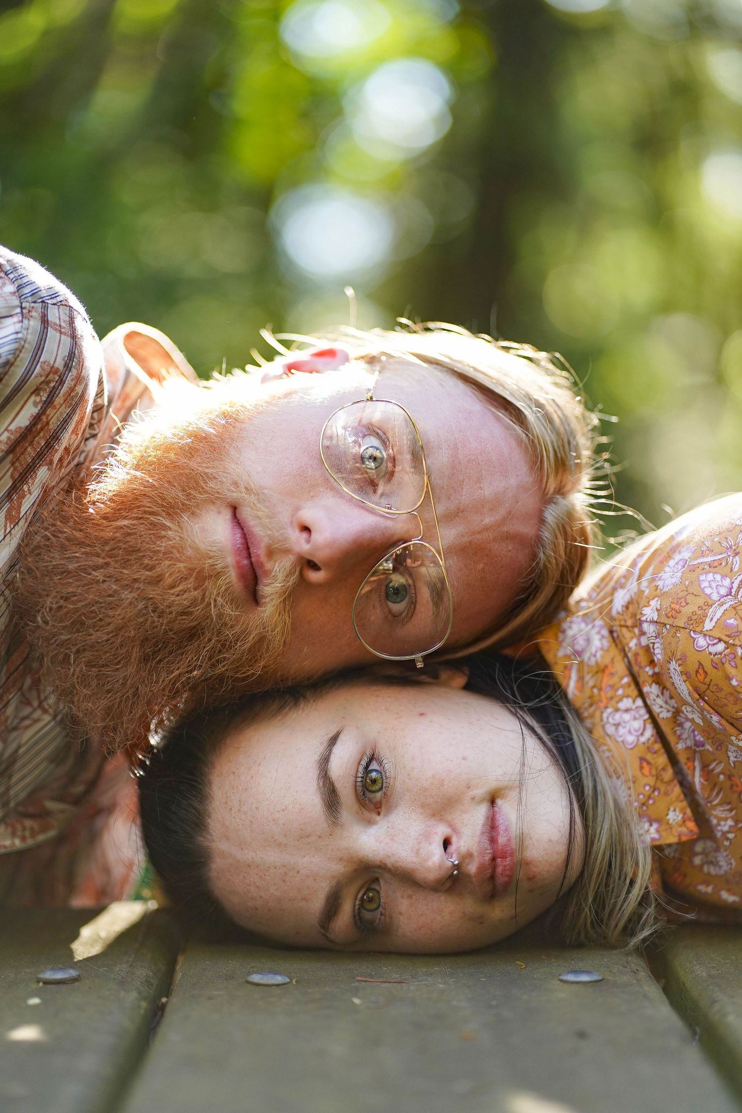 Two people lay head-to-head on wooden planks in a sunny forest setting.