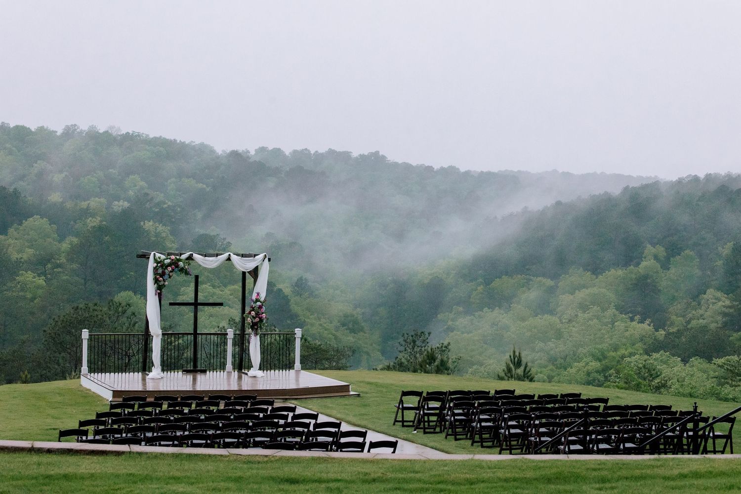 Elegant outdoor wedding ceremony setup with black chairs and floral arch overlooking misty forested hills.
