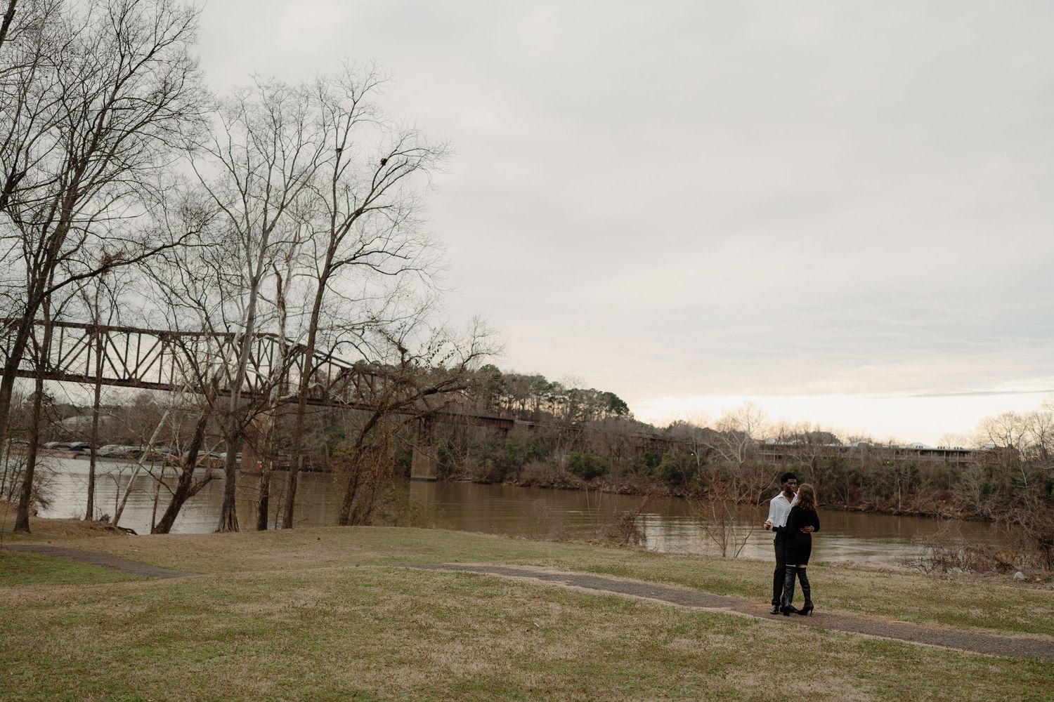 Couple embracing near a river with a historic iron bridge in the background at twilight.