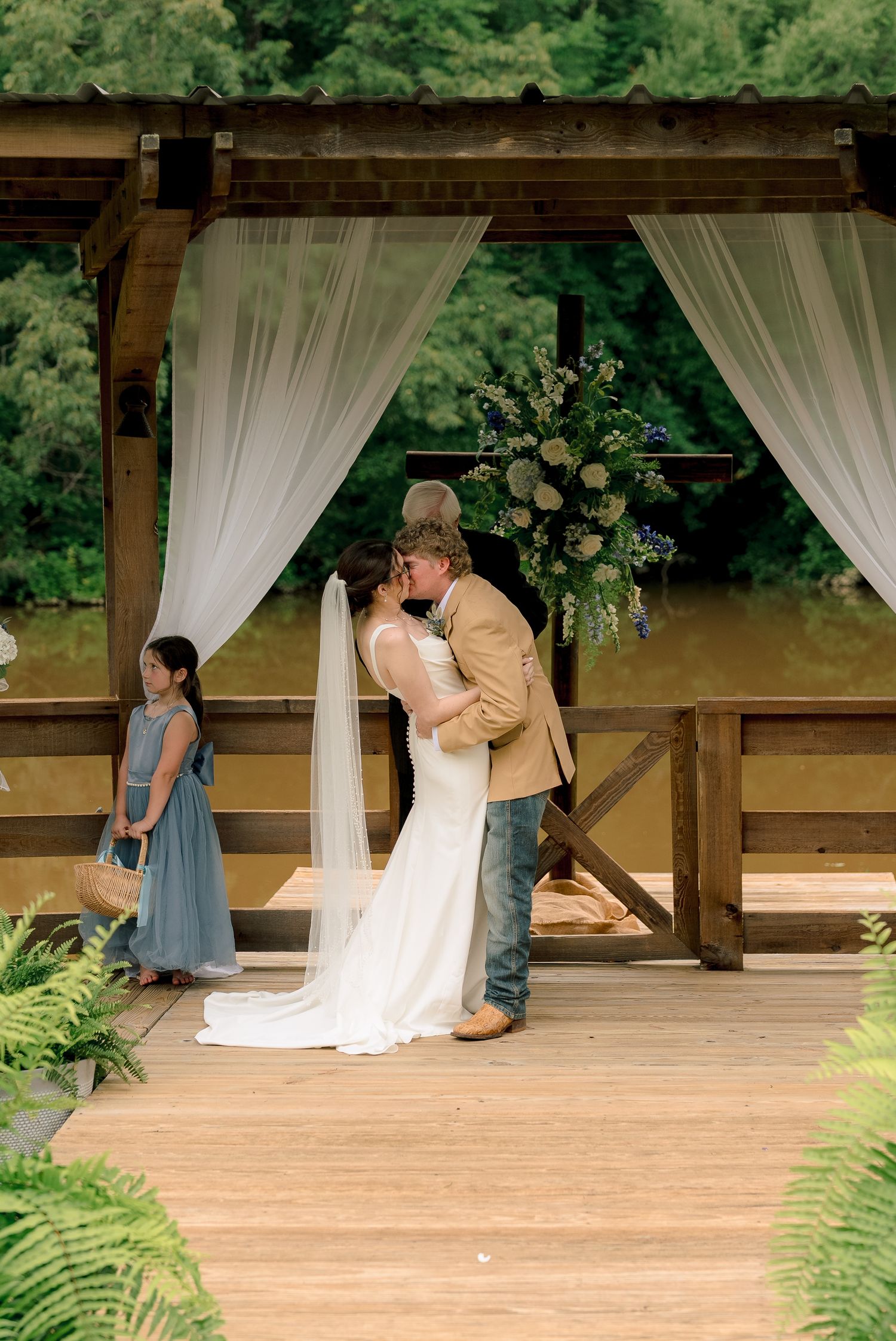 Bride and groom sharing first kiss under rustic wooden pergola with white drapes and floral arch.