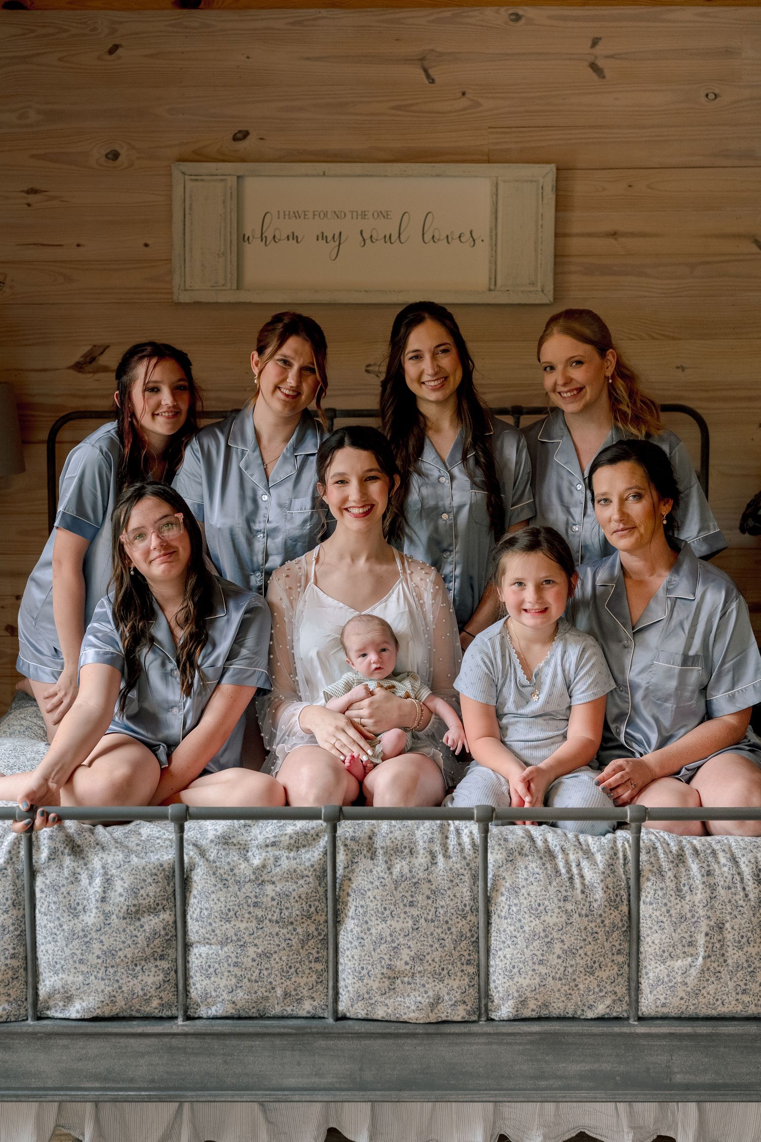 Bride in white lace robe holds newborn baby surrounded by bridesmaids in gray pajamas on rustic wooden bed.