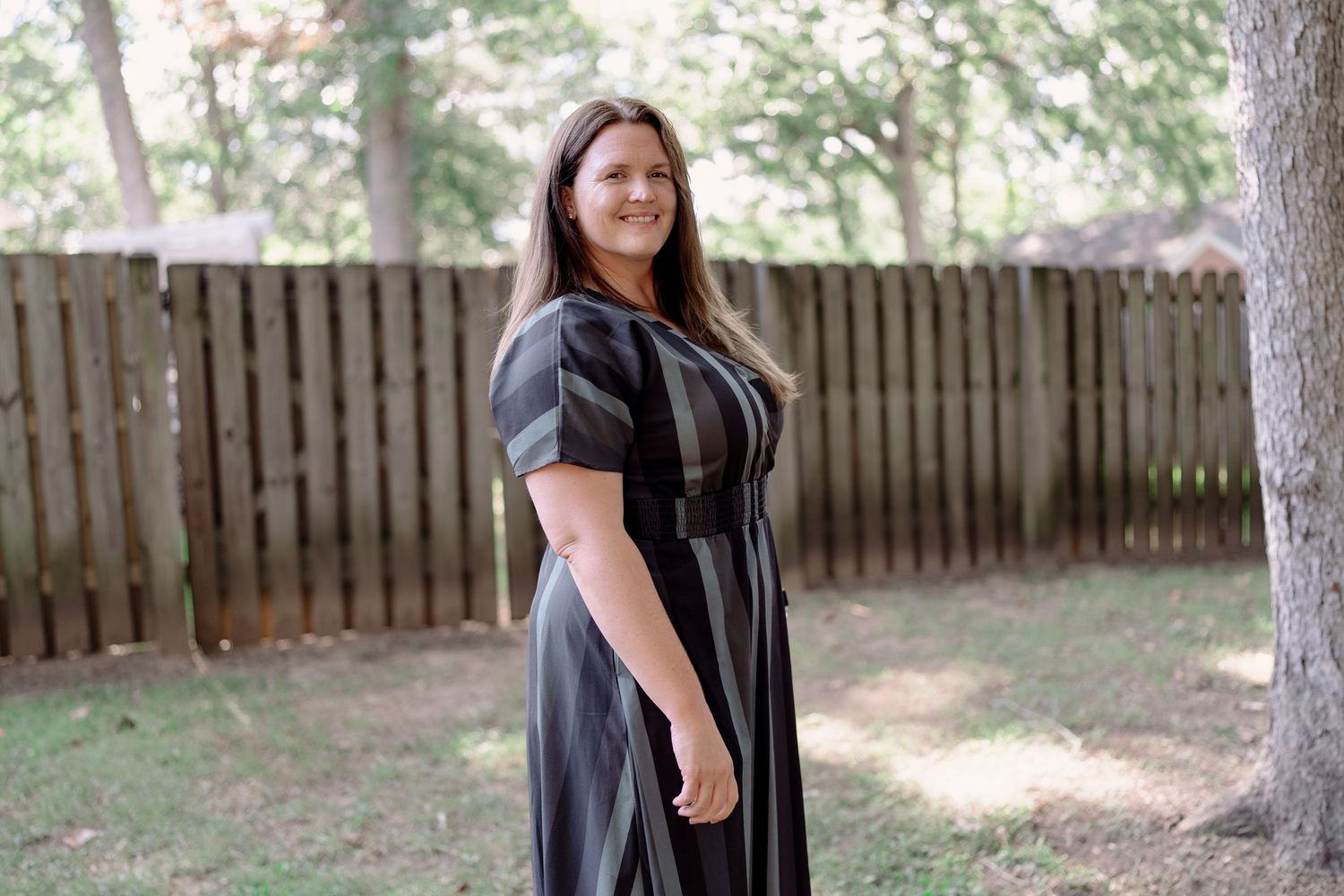 A woman in a black dress poses outdoors in a backyard setting with wooden fencing and trees in the background.