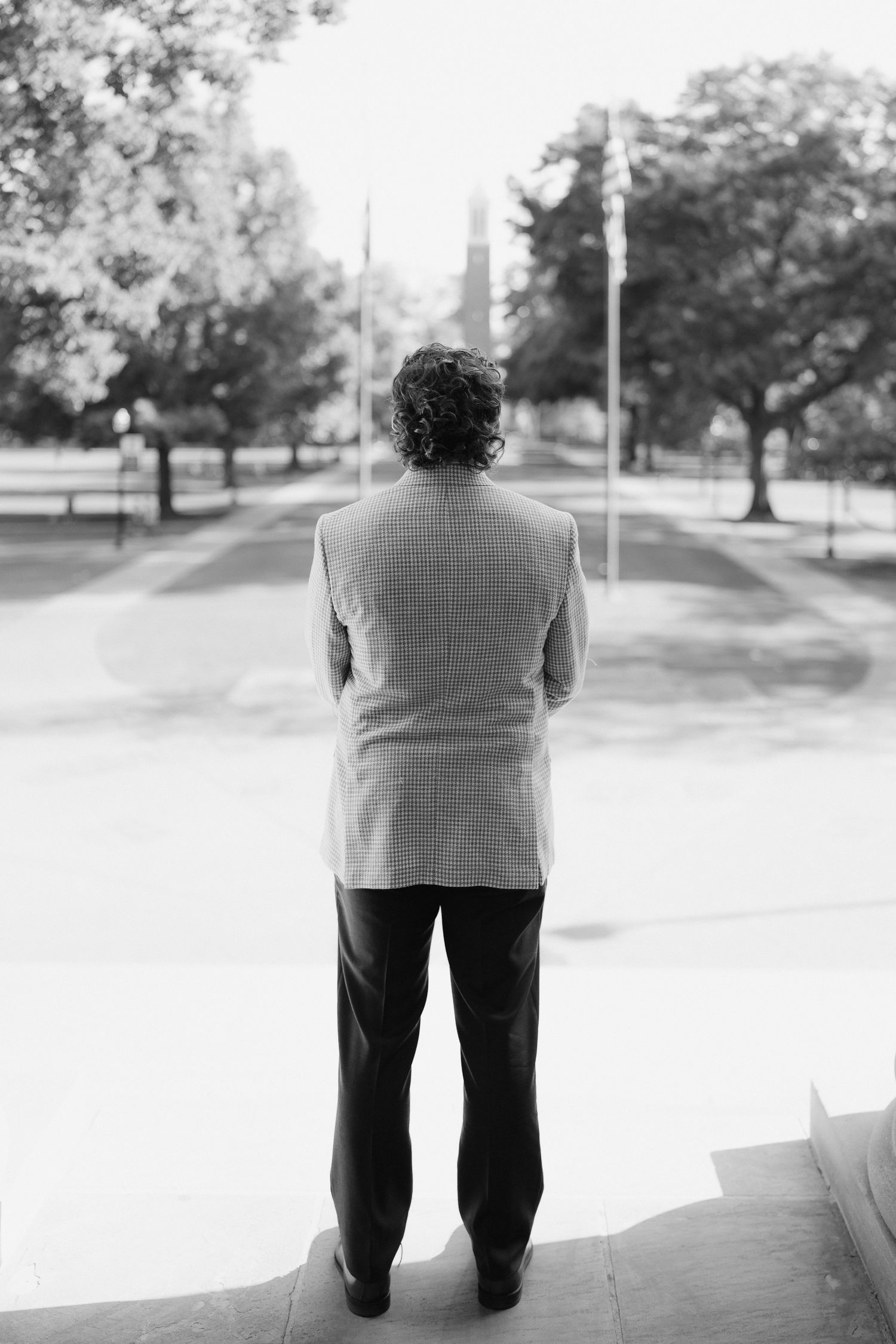 Man in gray blazer and dark trousers stands with back to camera overlooking a park with flagpoles.