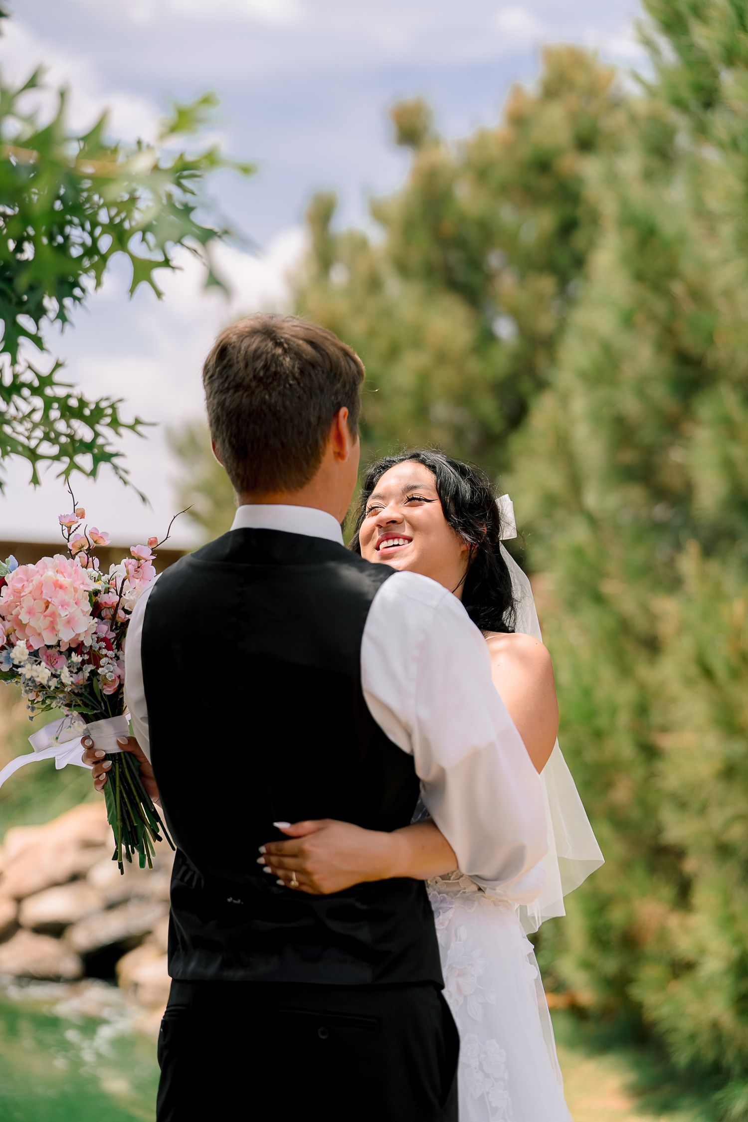 Smiling bride in white dress embraces groom in black vest outdoors with pink bouquet and green trees.