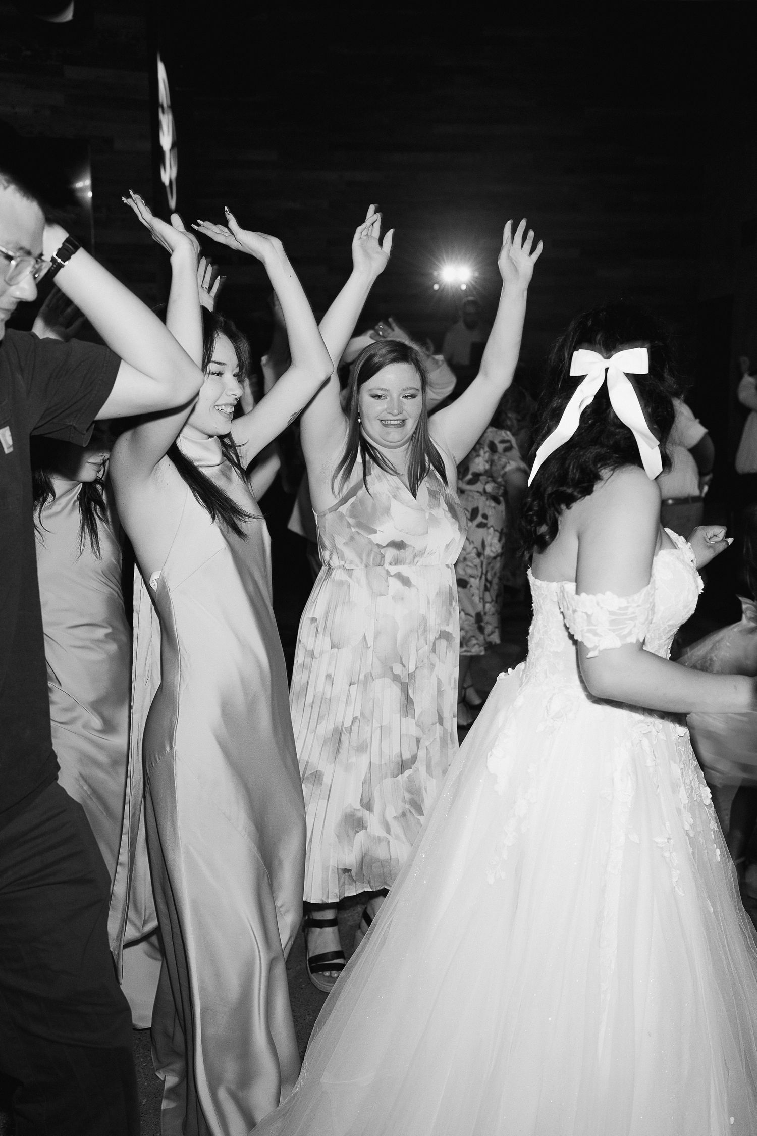 Black and white photo of wedding guests dancing joyfully, bride in white gown with bow visible at lively reception.
