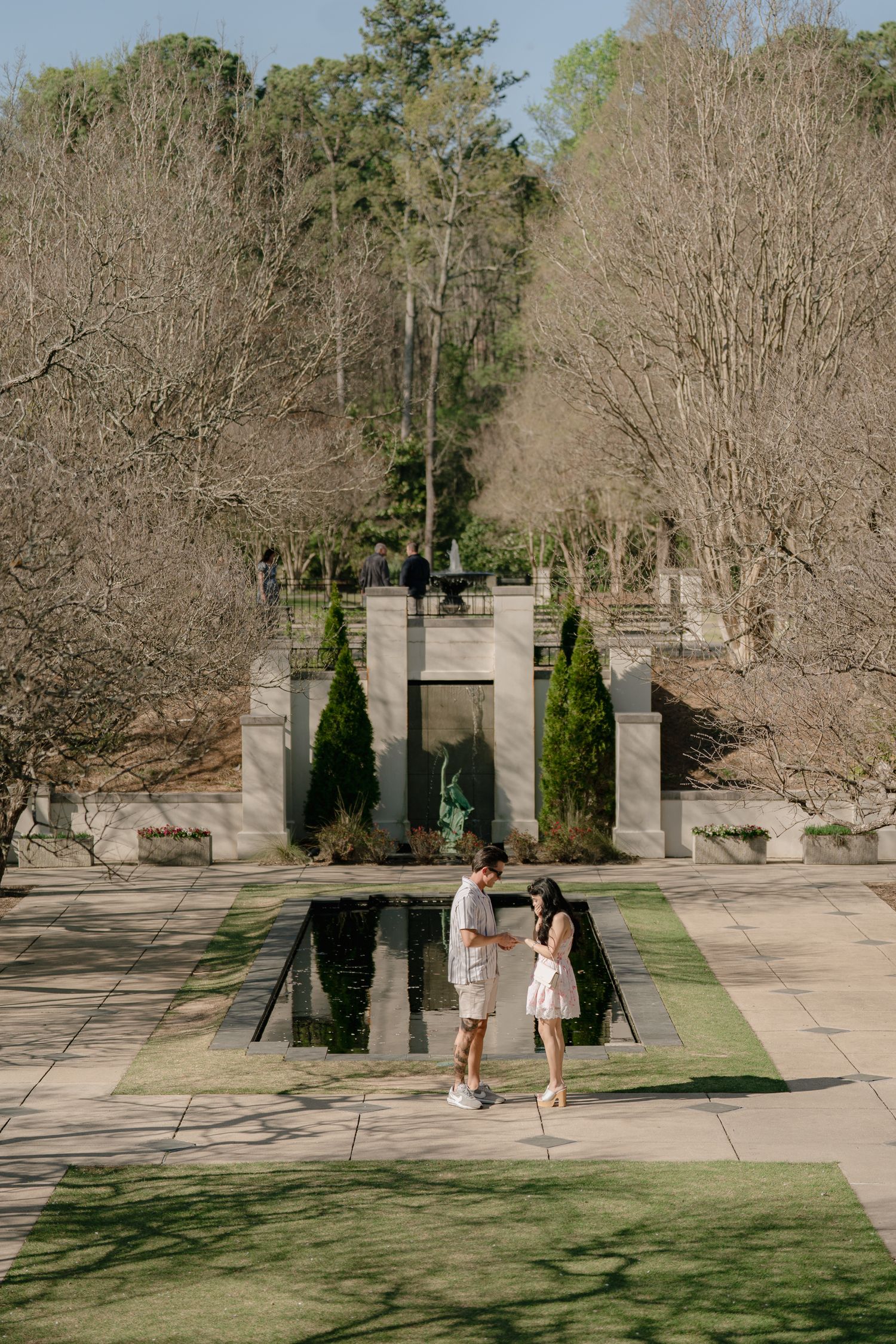 Couple standing by a reflective pool in a formal garden with stone architecture and cypress trees.