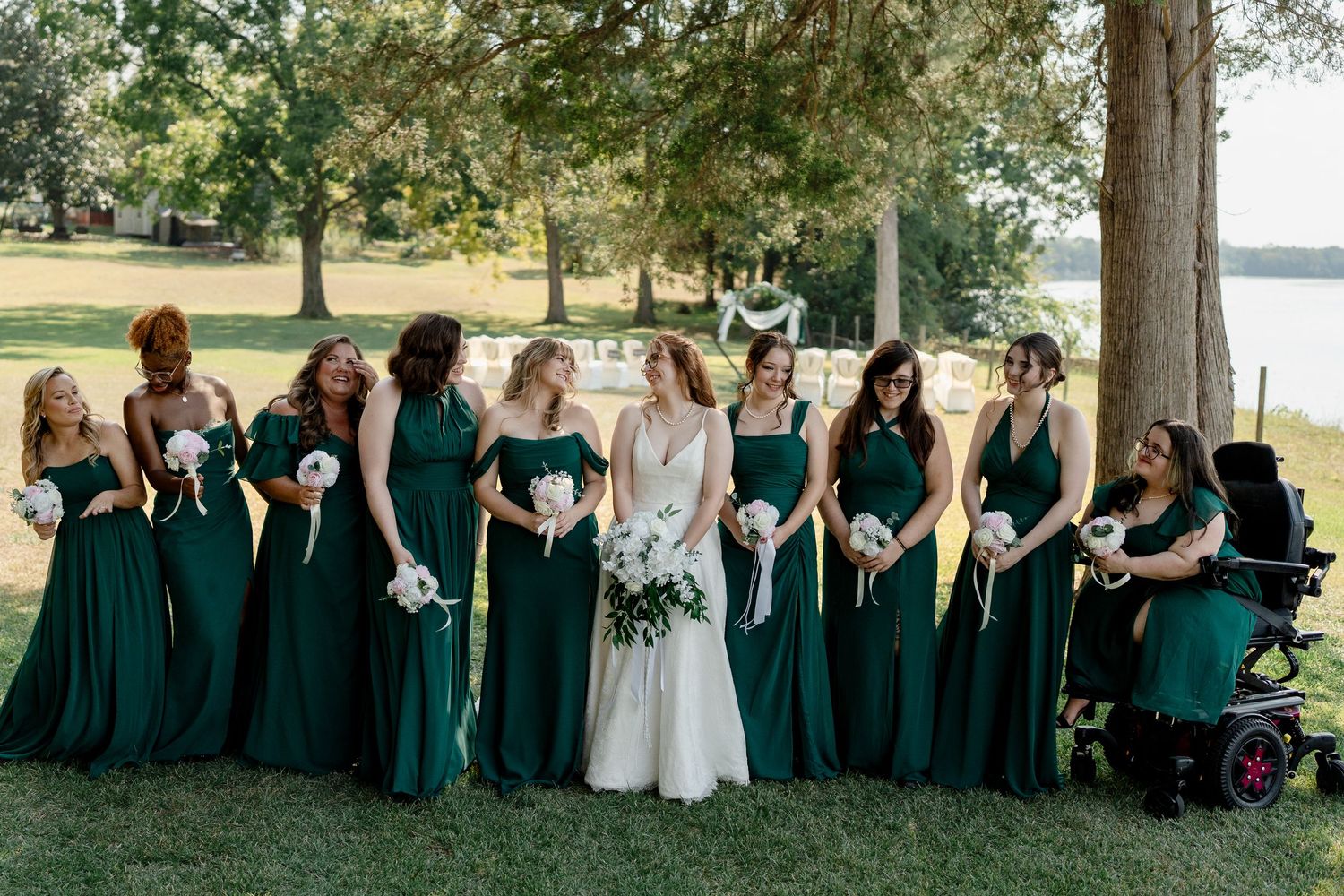 Wedding party in emerald green dresses and black suits poses outdoors on a sunny day by the lake.