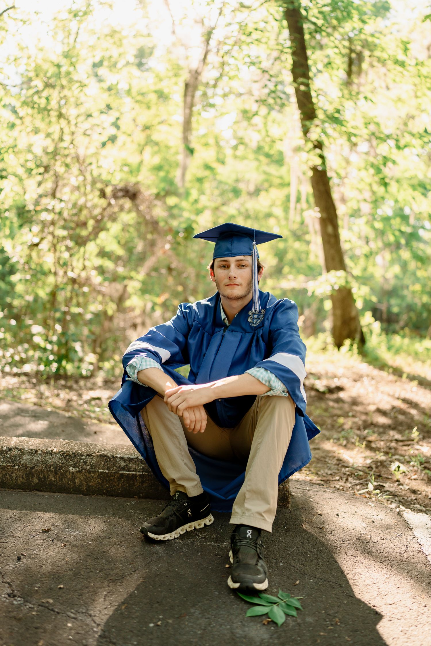 A graduate in academic regalia sits casually on a path in a wooded setting.
