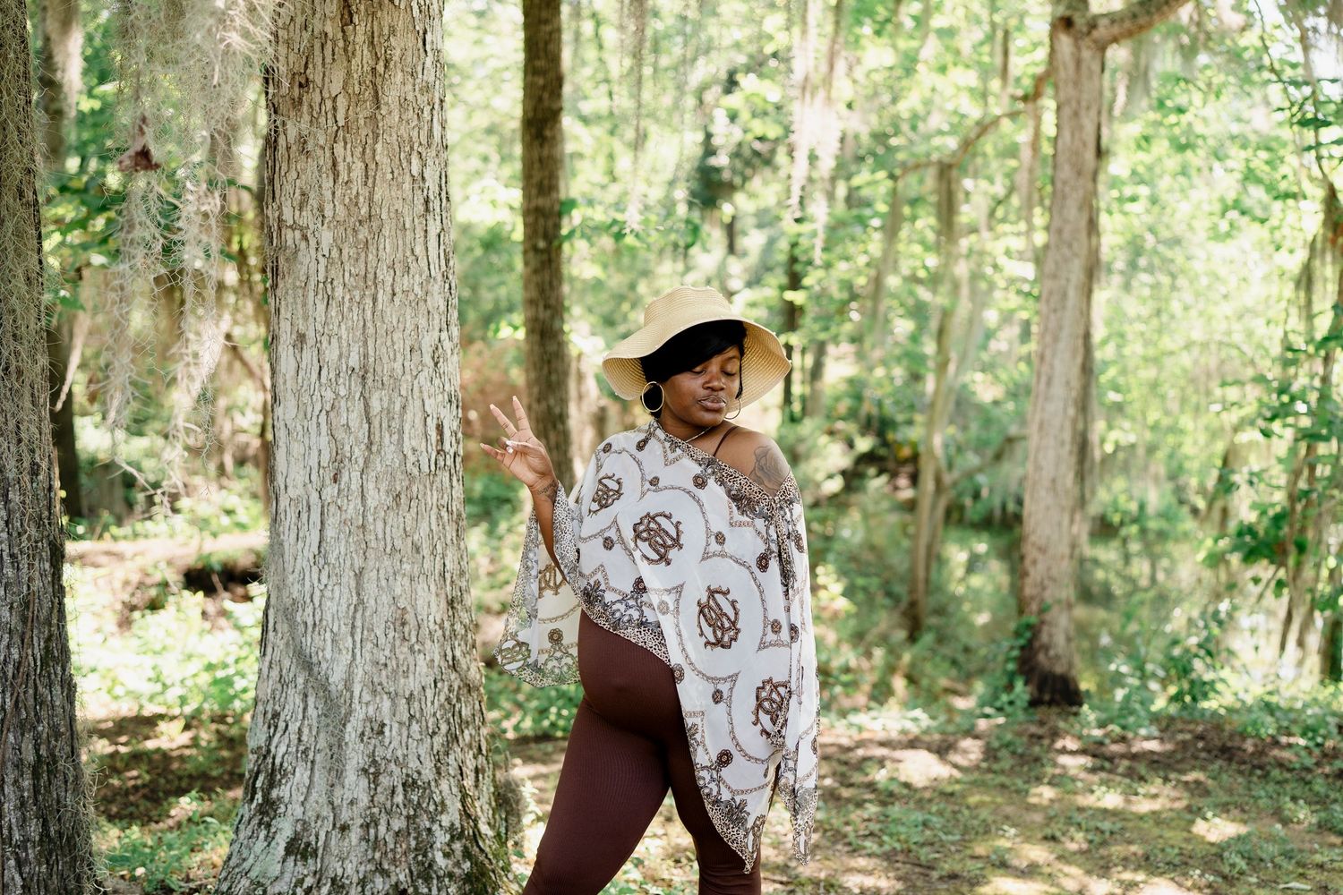 Person in patterned white shawl and wide-brimmed hat poses playfully by a tree trunk.