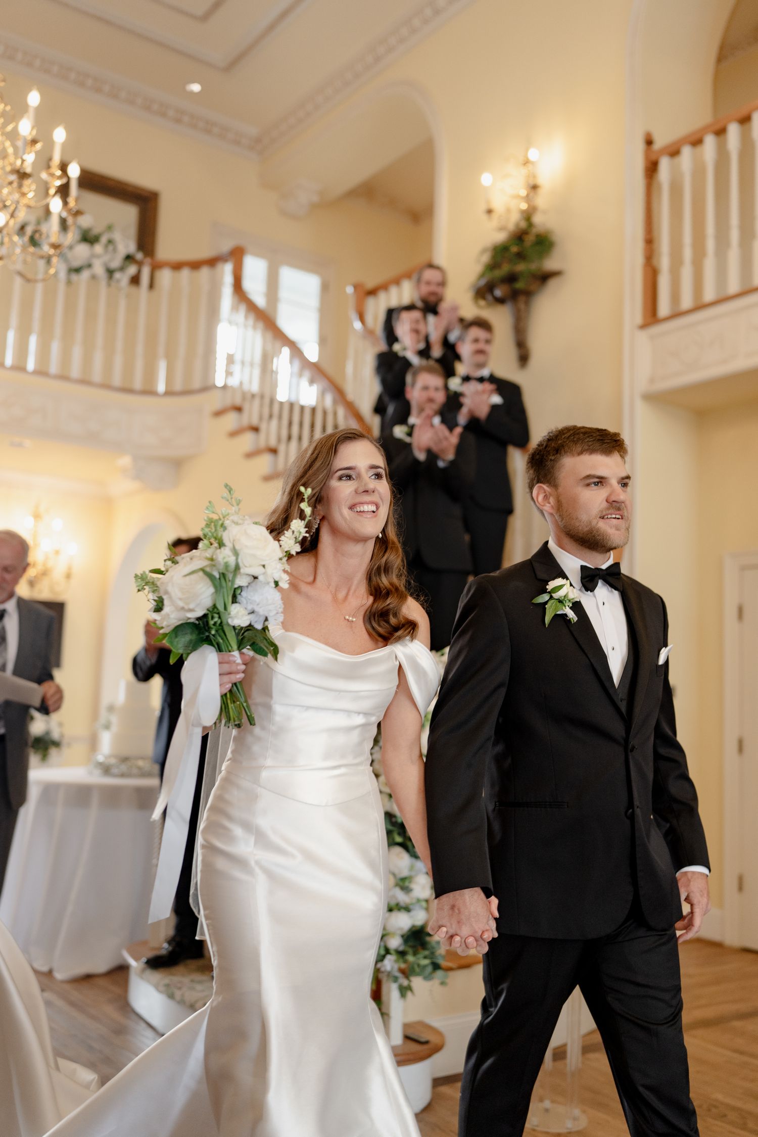 Joyful wedding couple walks together down aisle in elegant venue with grand staircase and chandelier.