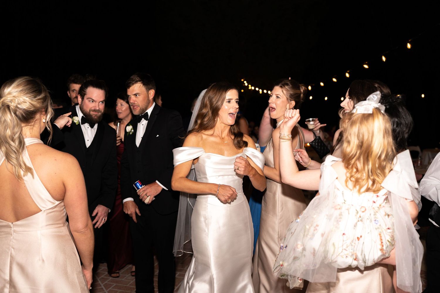 Wedding reception celebration with people in white dresses dancing under string lights.