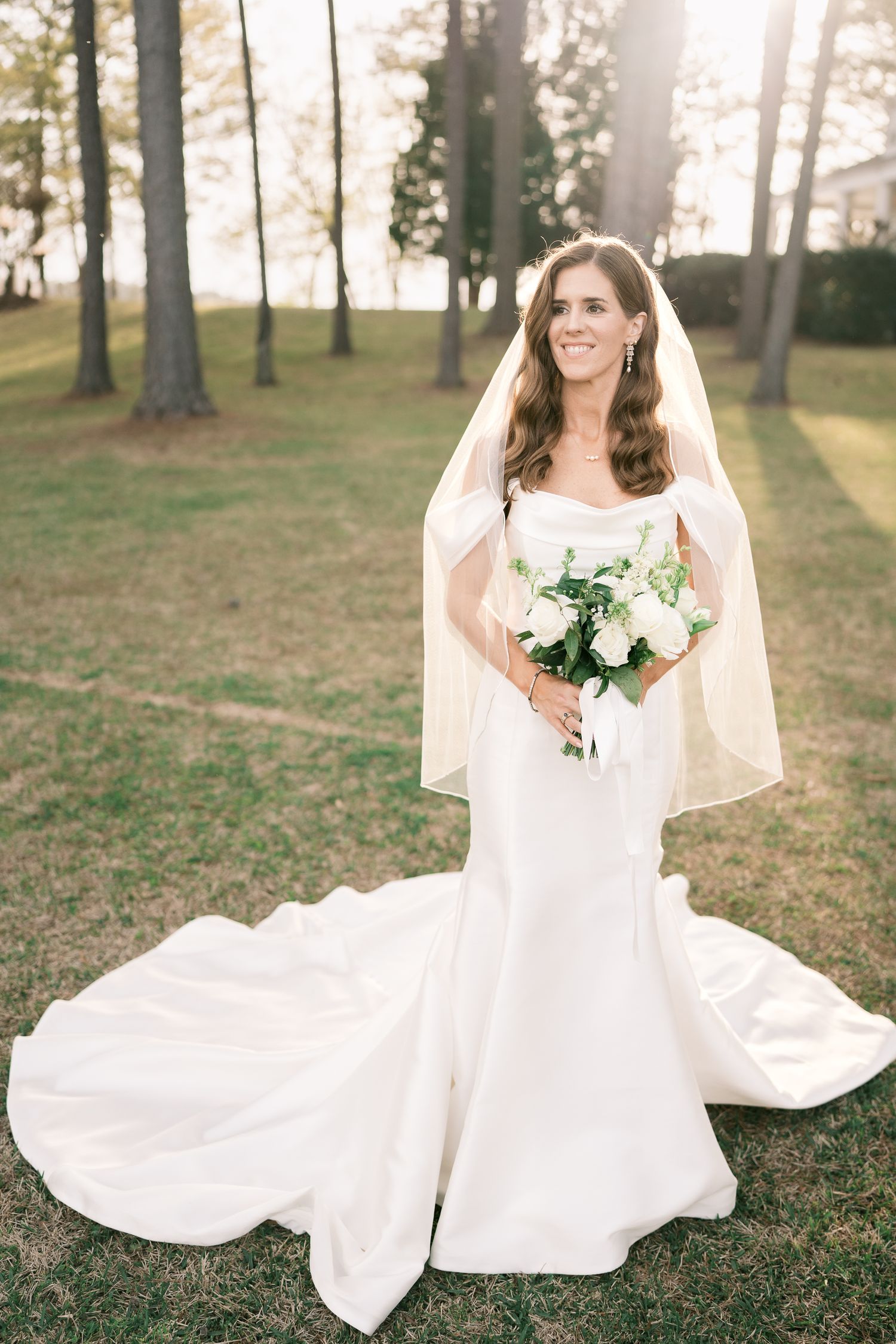 Bride in white mermaid gown with long veil poses with bouquet on sunny lawn.