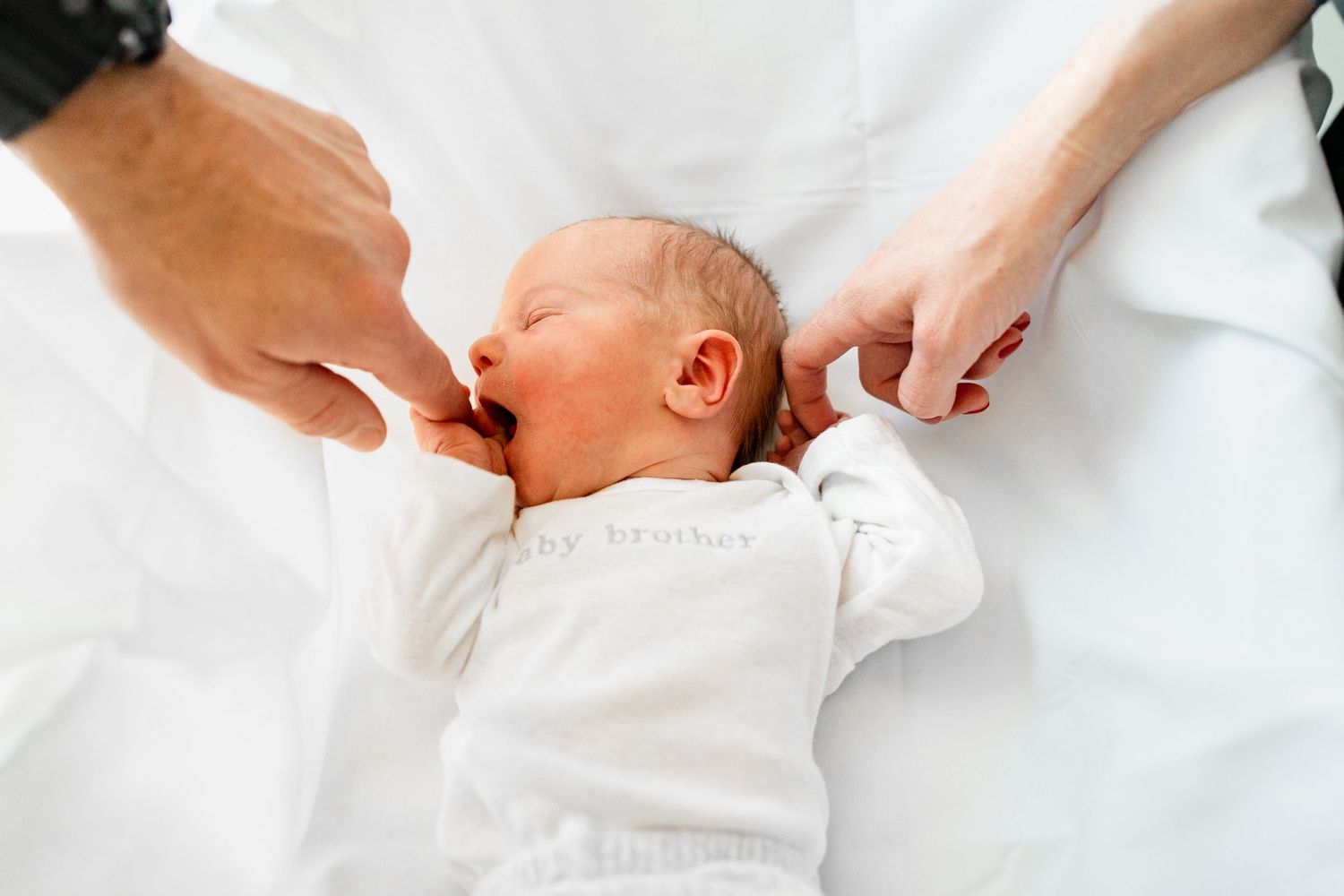 Newborn in white outfit resting on clean white bedding with caring hands nearby.