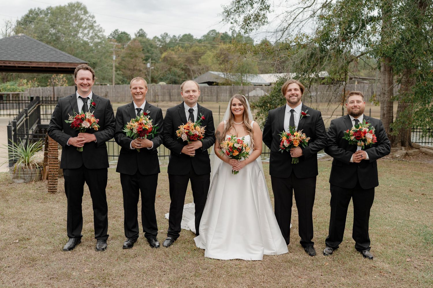 Wedding party stands in a row outdoors with groomsmen in black suits and bridesmaids holding orange bouquets.