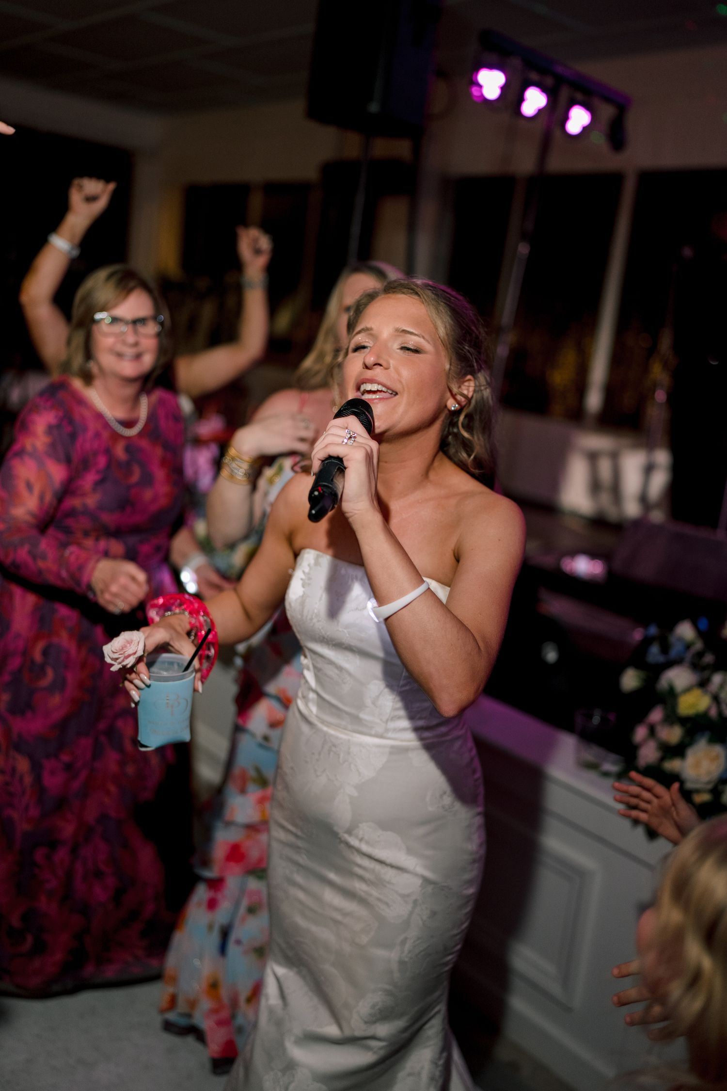 Bride in white gown singing into microphone at wedding reception with guests dancing in background.