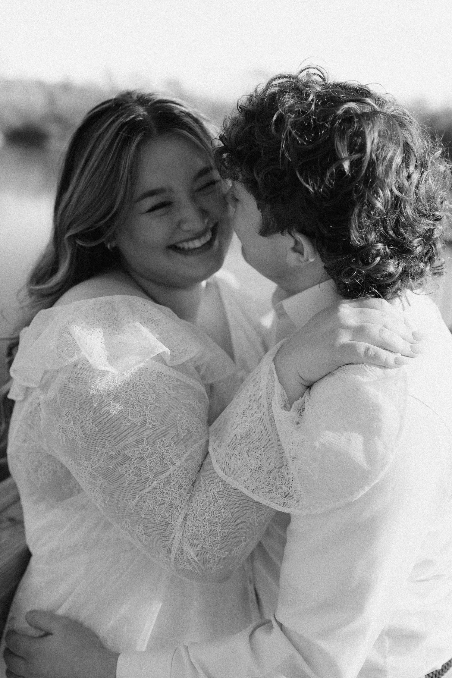 Black and white close-up of laughing woman embracing curly-haired man near a blurred lake background.