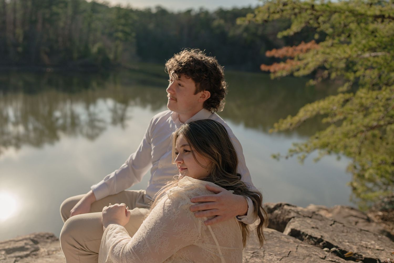 A couple sits on rocks by a serene lake surrounded by lush green trees in soft natural light.