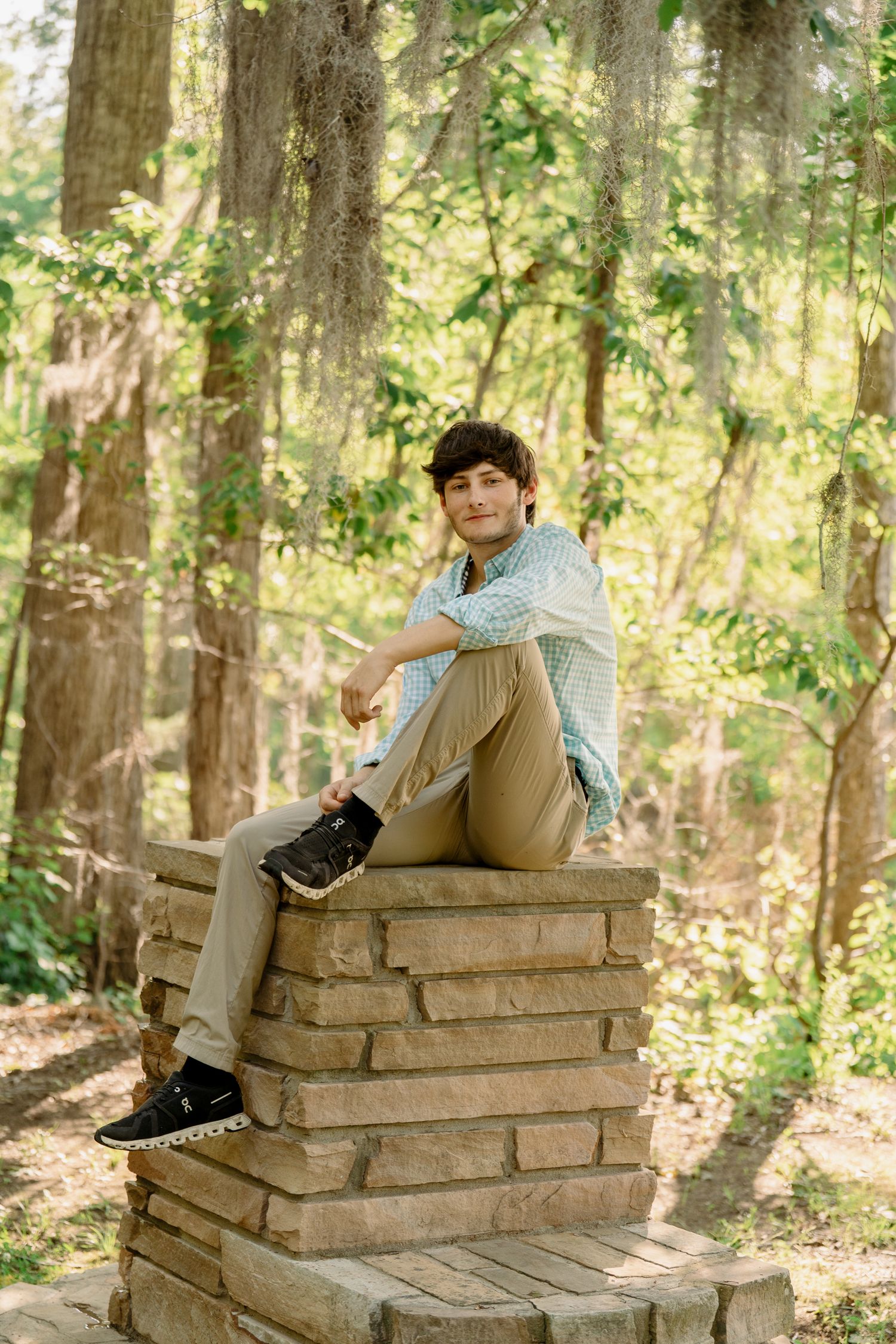 Graduate in casual attire sits on stone structure in sunlit wooded area.
