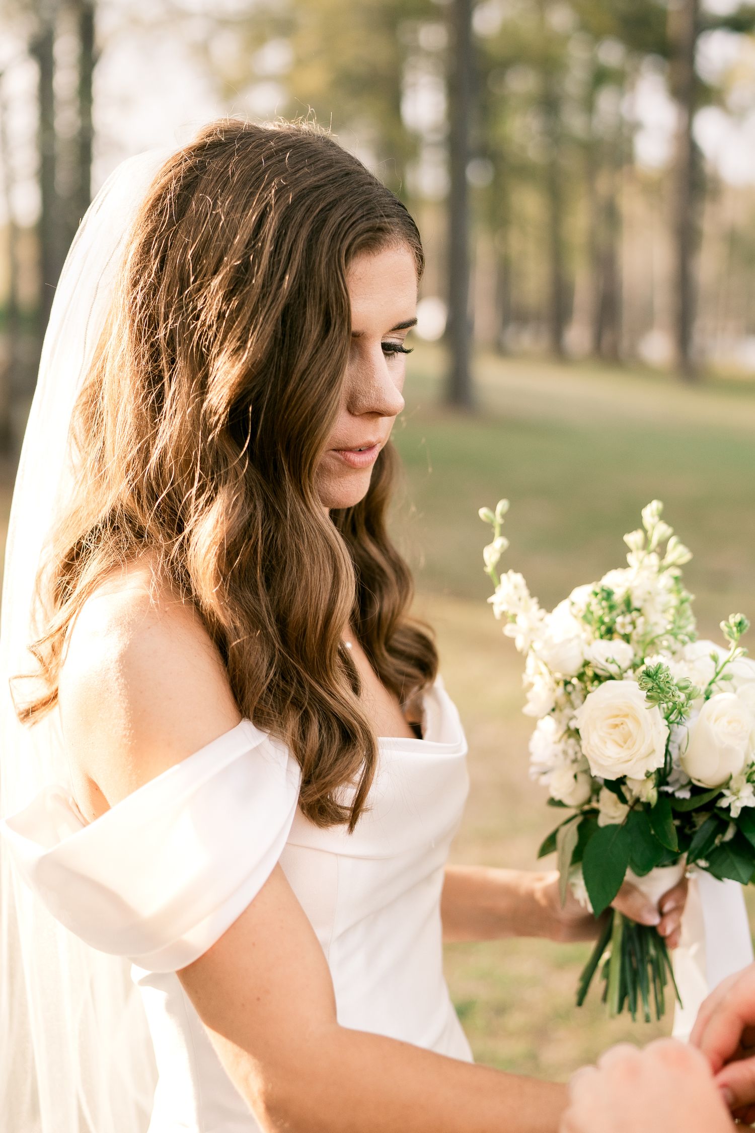 Elegant bride in white off-shoulder dress holding a delicate white and green bouquet in a sunlit forest setting.