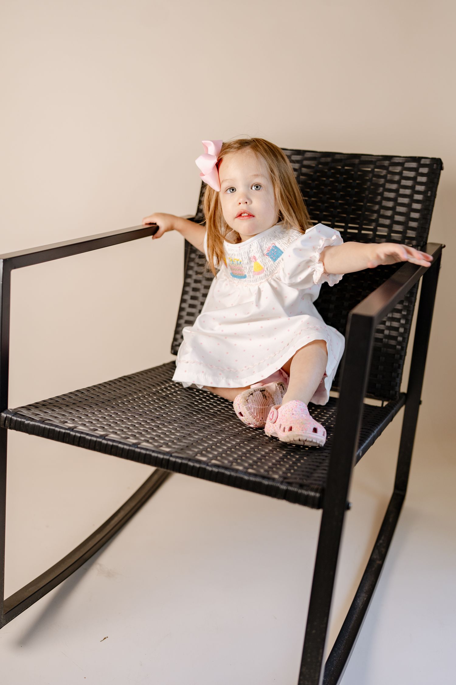Child in white dress poses playfully on modern black chair.
