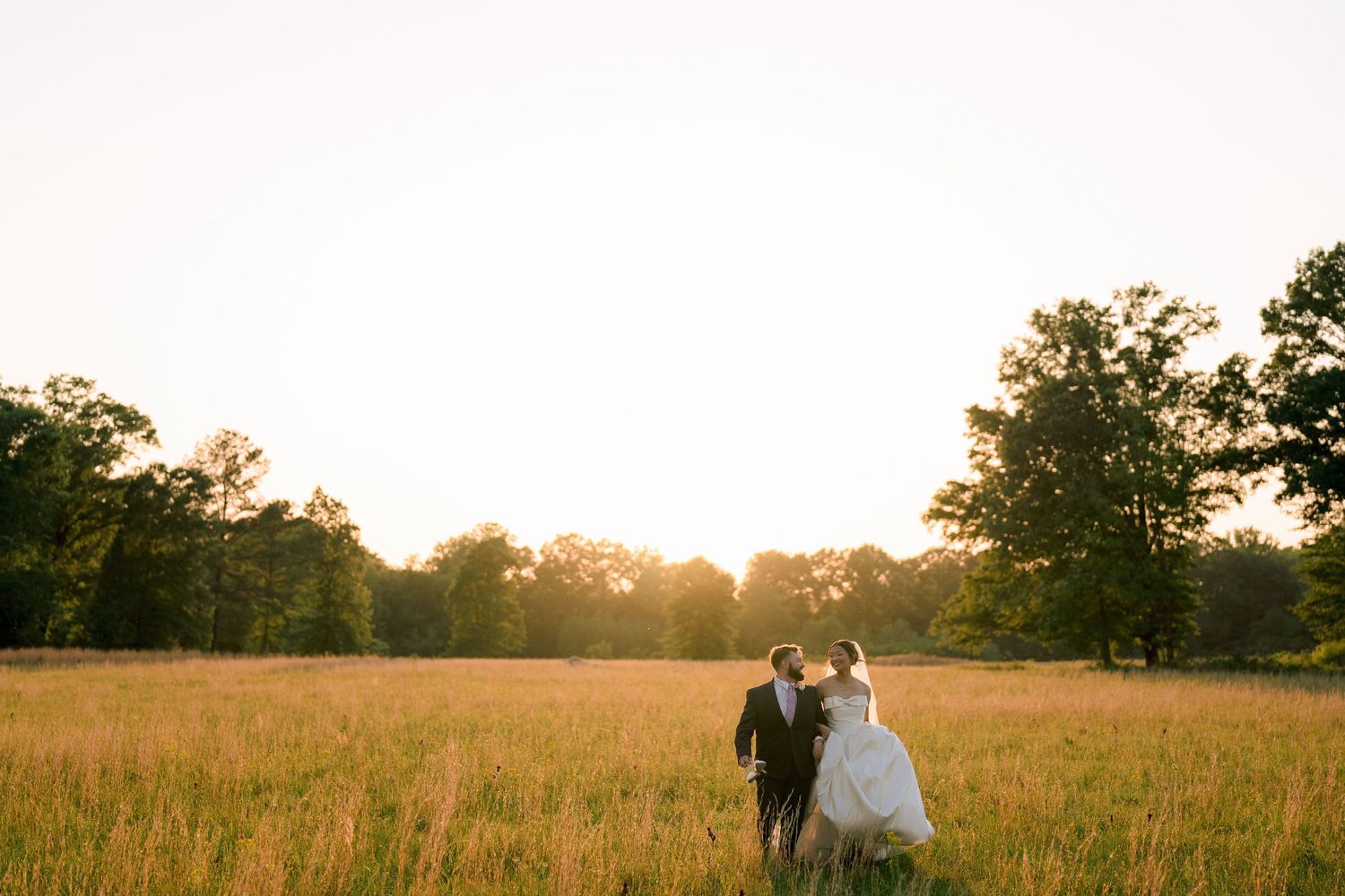 Bride and groom walking hand in hand through golden sunlit field at sunset with trees in background.