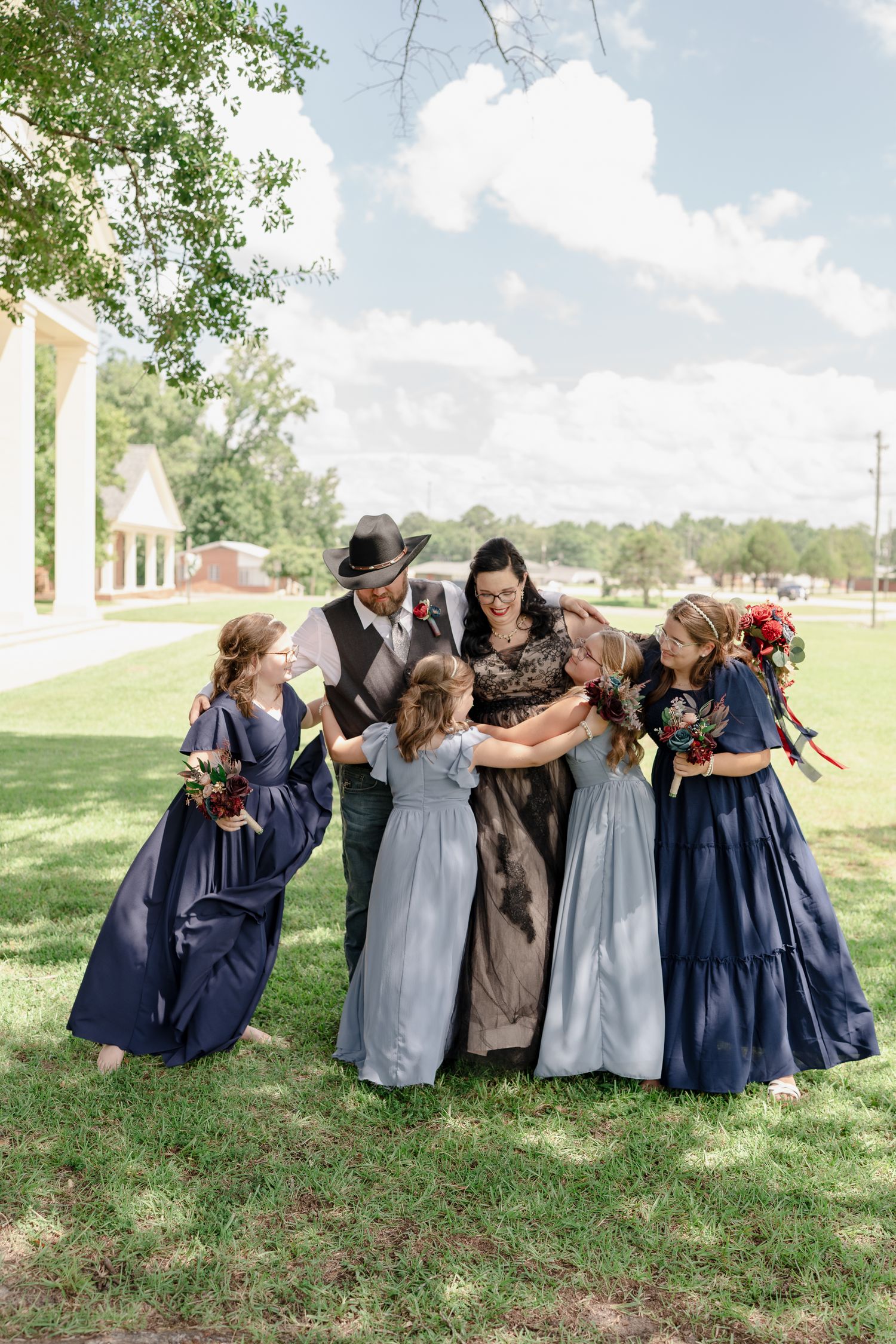 A group in vintage-style dresses huddles together laughing on a sunny lawn with trees and white columns in the background.
