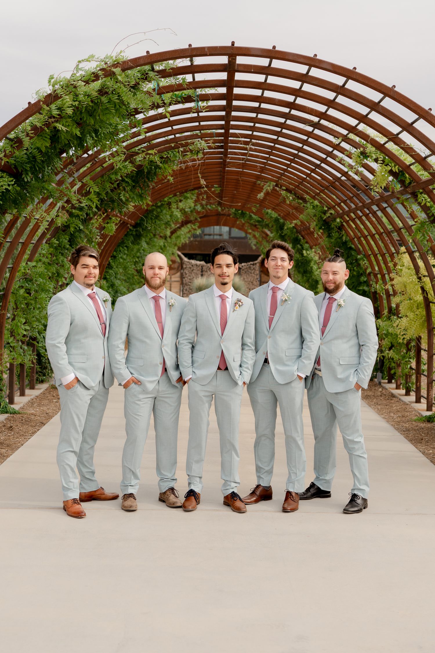 Groomsmen in light gray suits with coral ties pose together under a wooden pergola at an outdoor venue.