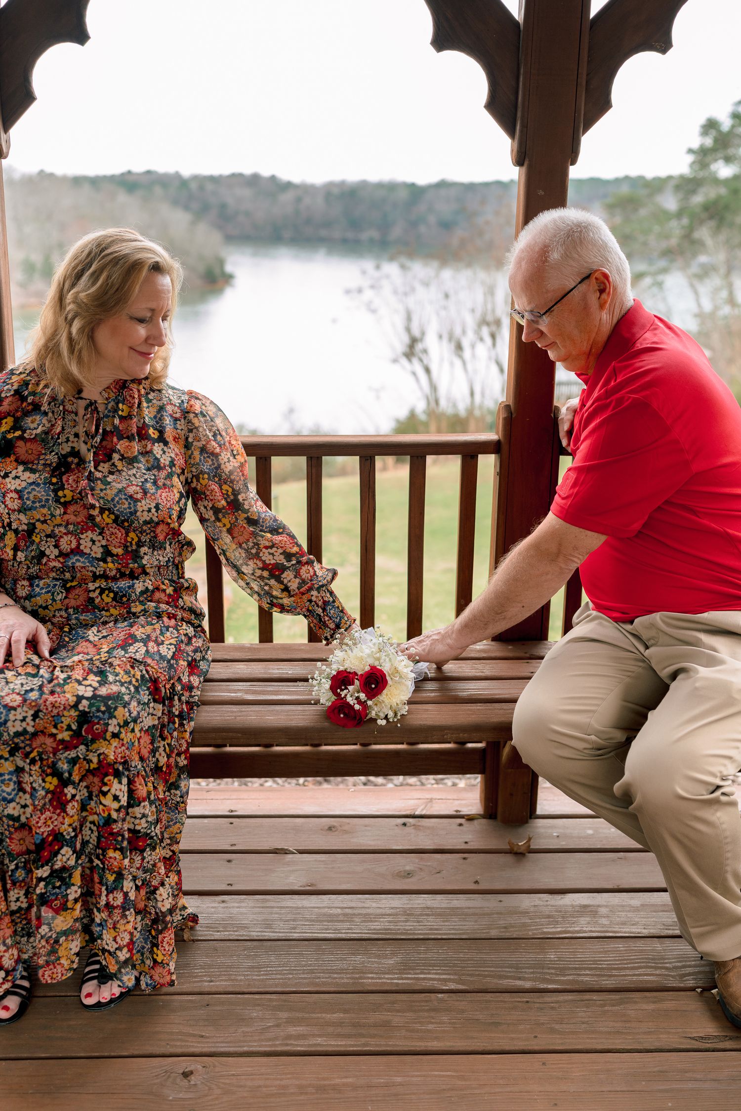Older man presenting a floral bouquet to a smiling woman seated on a bench in a wooden gazebo.