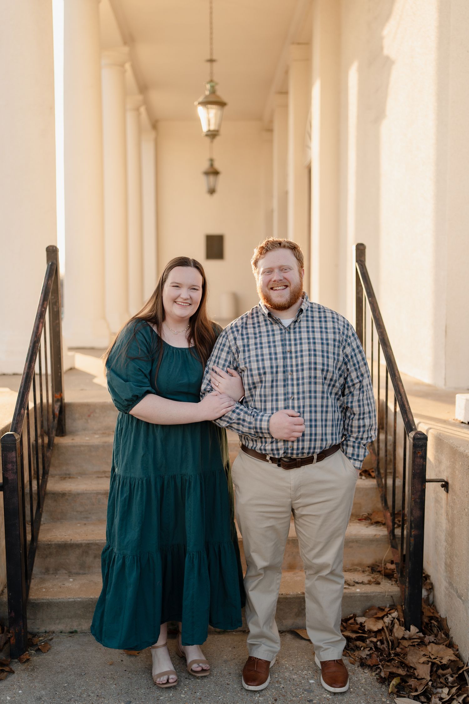 Couple in green dress and plaid shirt smiling arm-in-arm in a sunlit columned hallway.