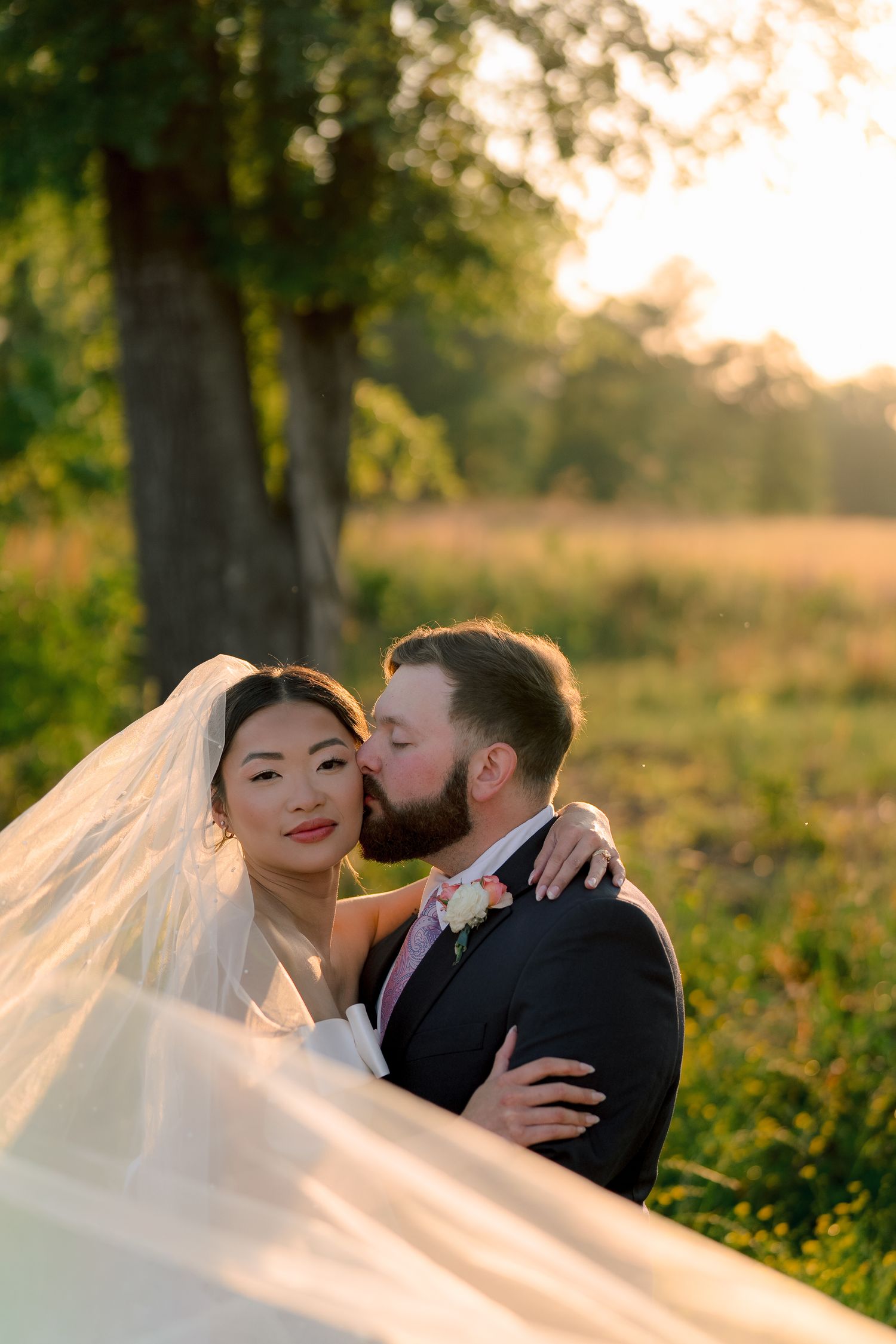 Bride and groom share a romantic kiss at golden hour, with veil flowing, in a sunlit outdoor field setting.