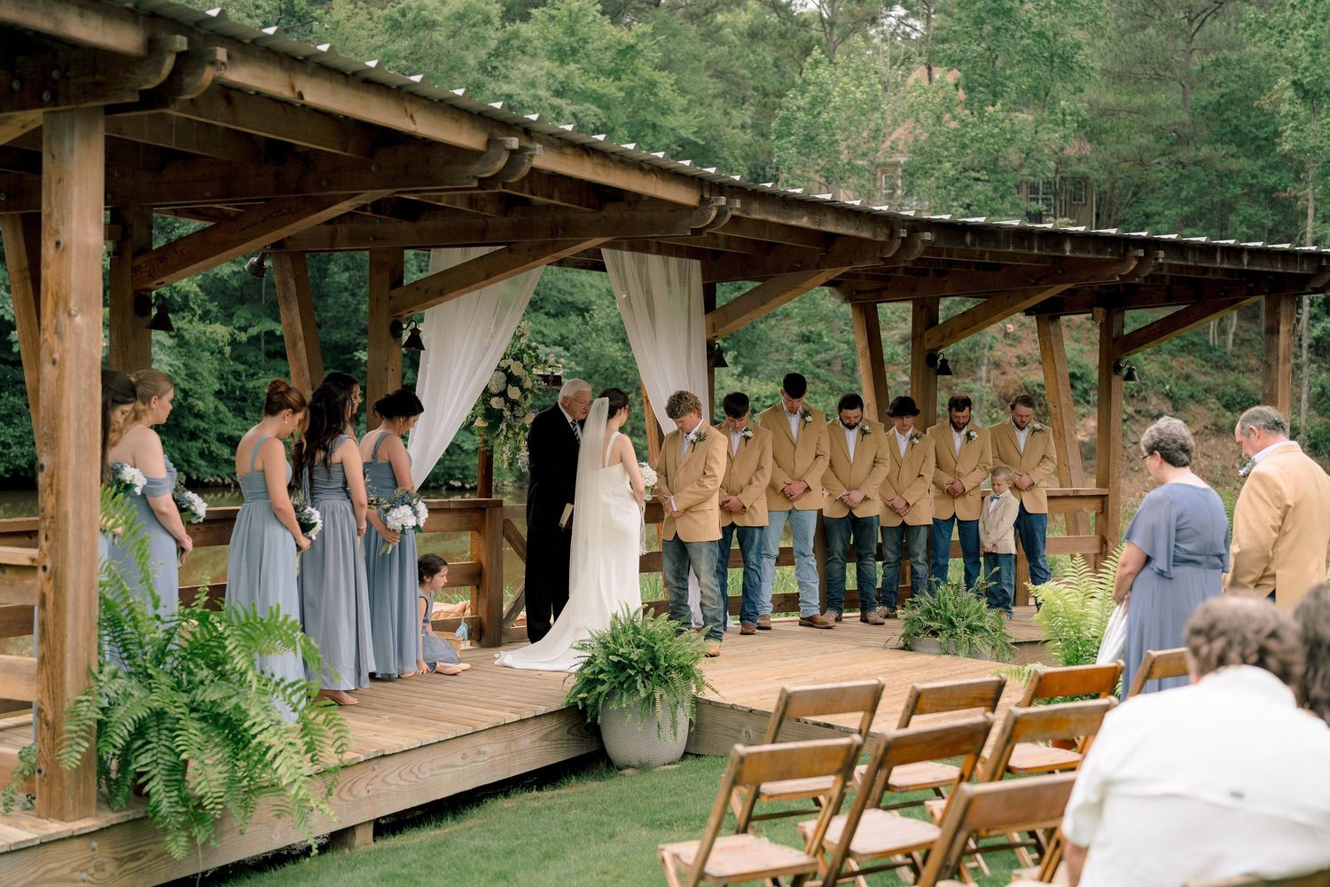 Outdoor wedding ceremony under wooden pergola with bridesmaids in blue-gray gowns and groomsmen in tan jackets.