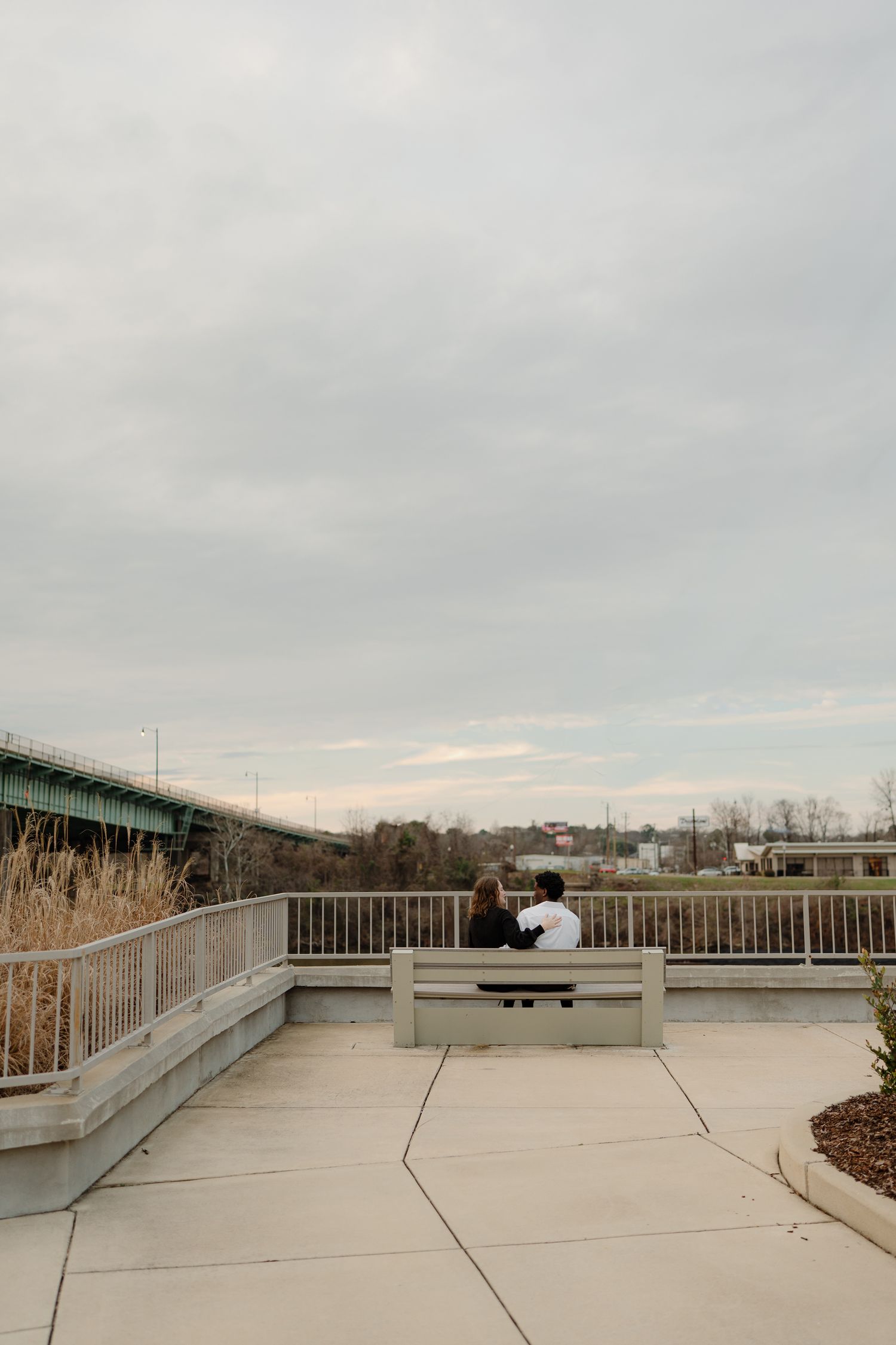 Couple sitting together on outdoor bench overlooking river and bridge under cloudy sky.