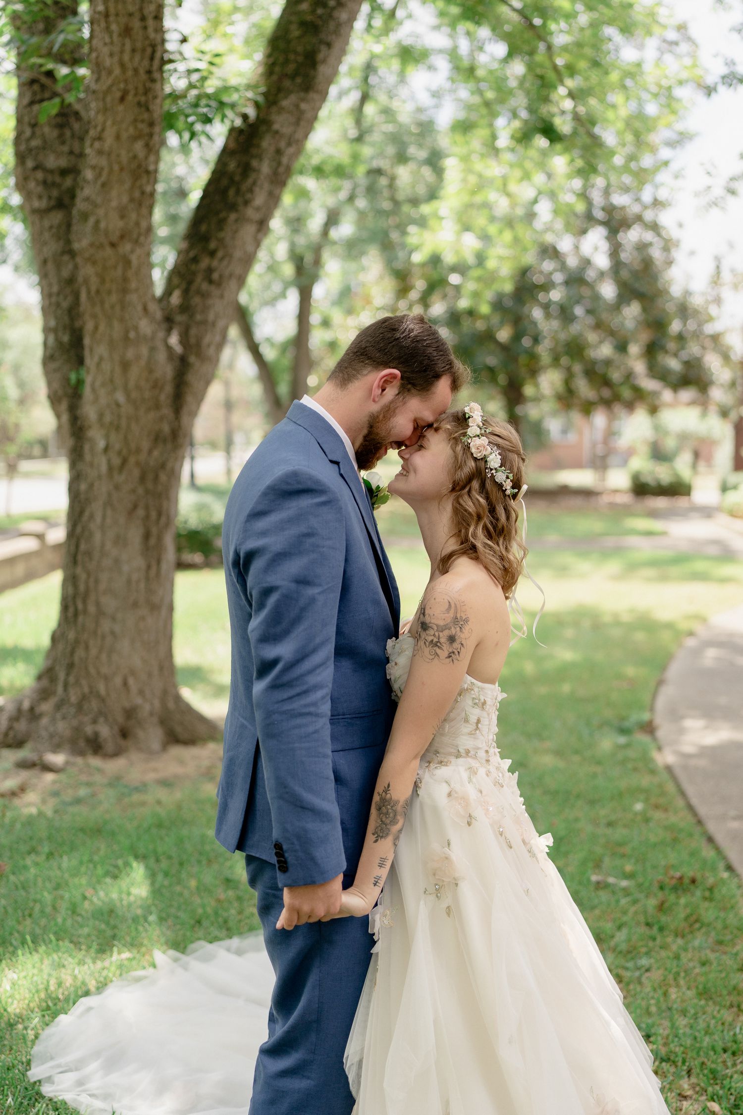 A newlywed couple embraces and dances joyfully along a tree-lined path in a park setting.