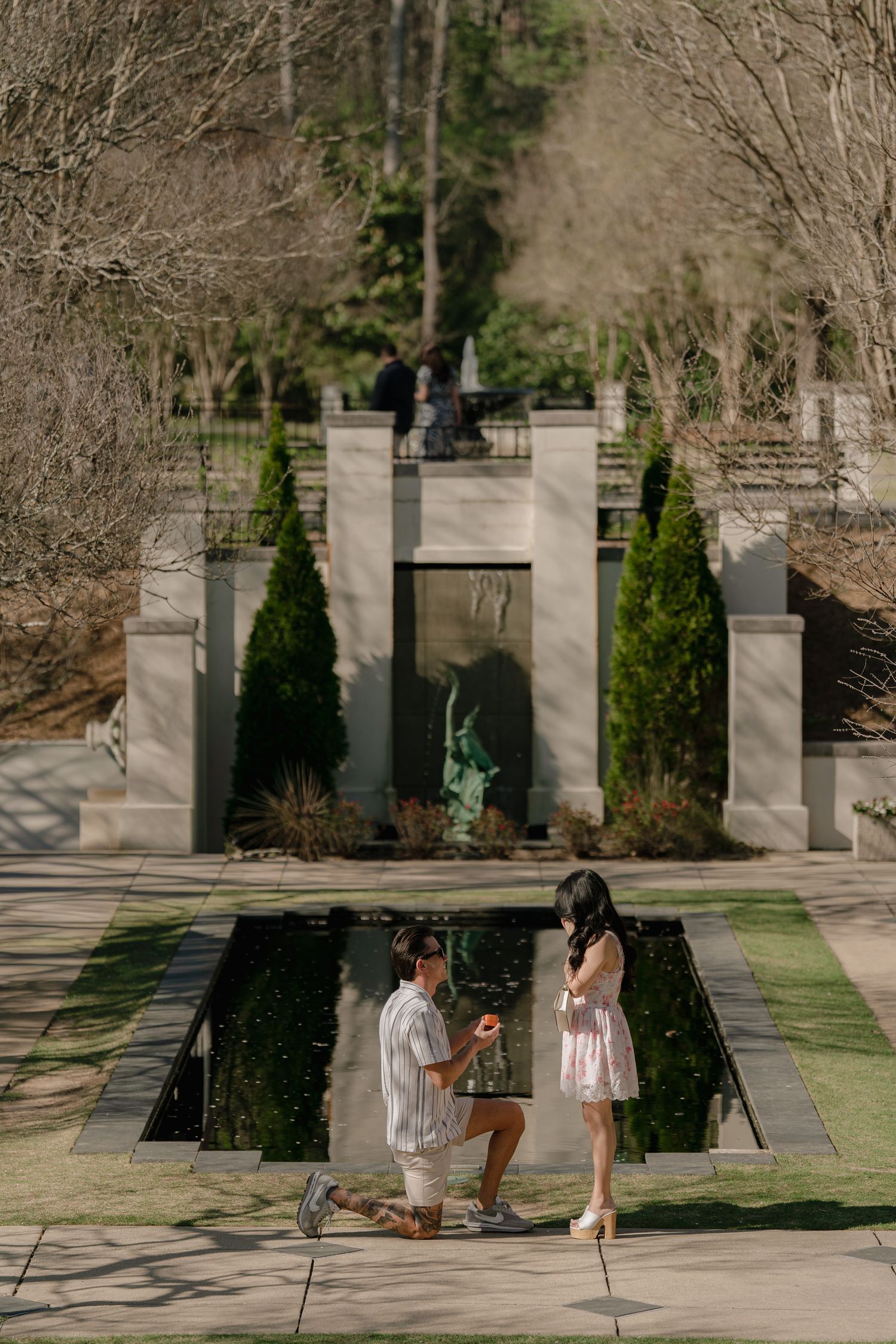 Man proposes on one knee to woman by a reflecting pool in an elegant garden with stone fountain.