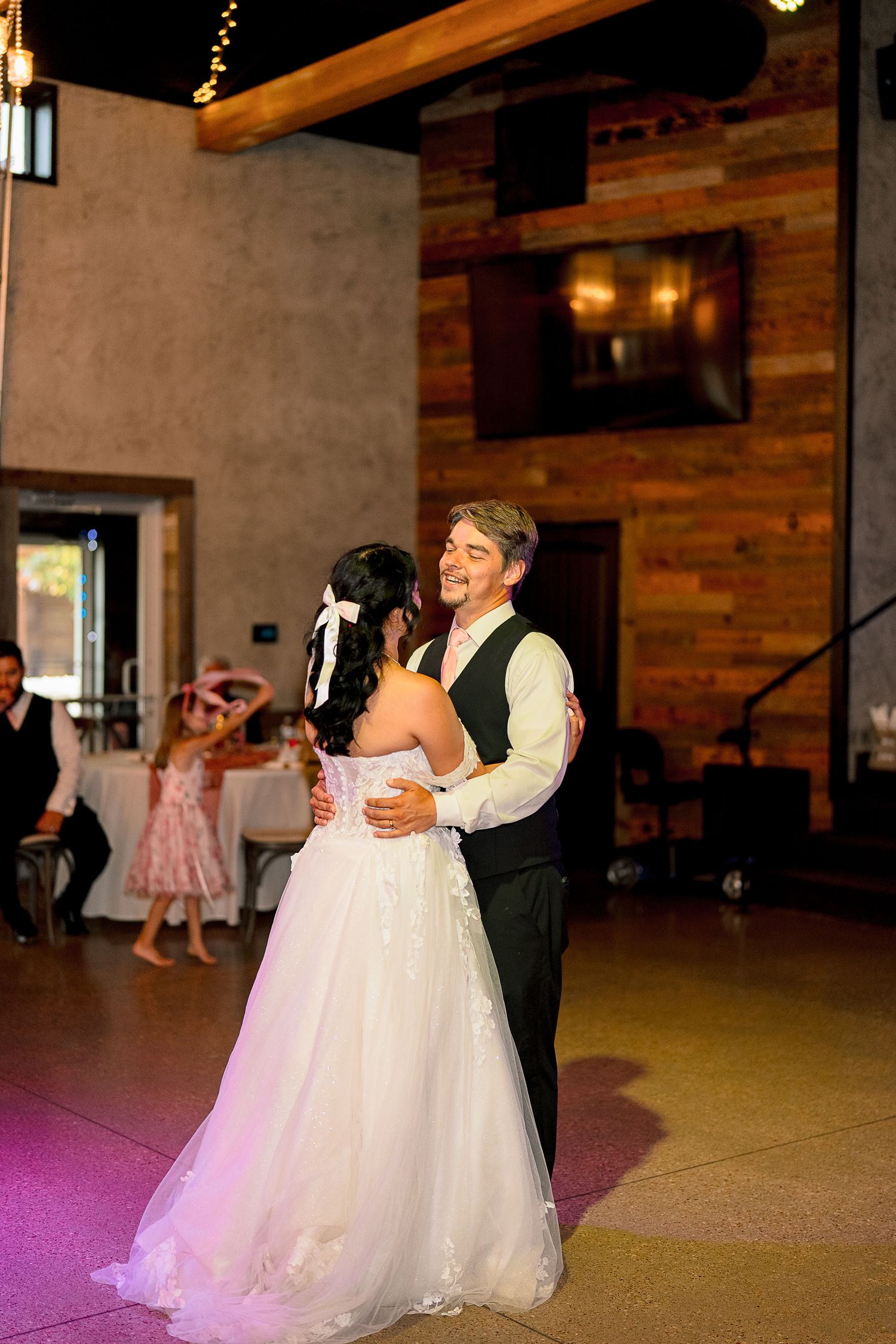 Bride and groom share first dance in rustic wedding reception venue with warm lighting and wood accents.