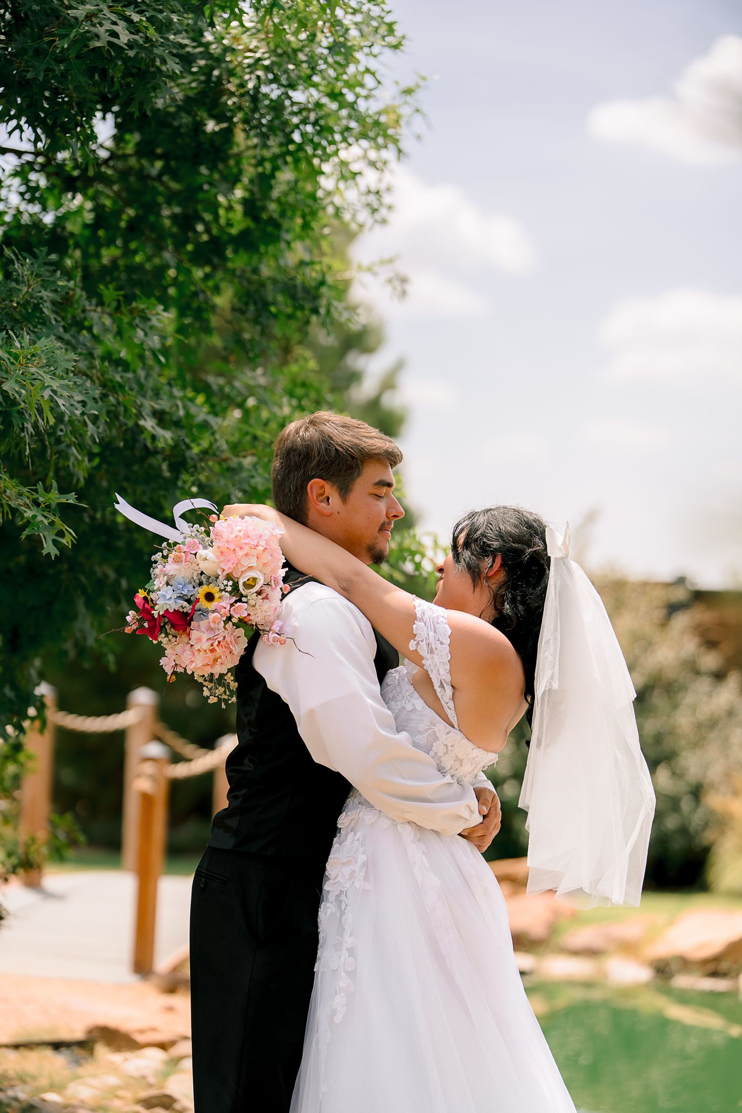 A bride and groom embrace outdoors, she holds a pink bouquet, her veil flowing in the breeze on a sunny day.