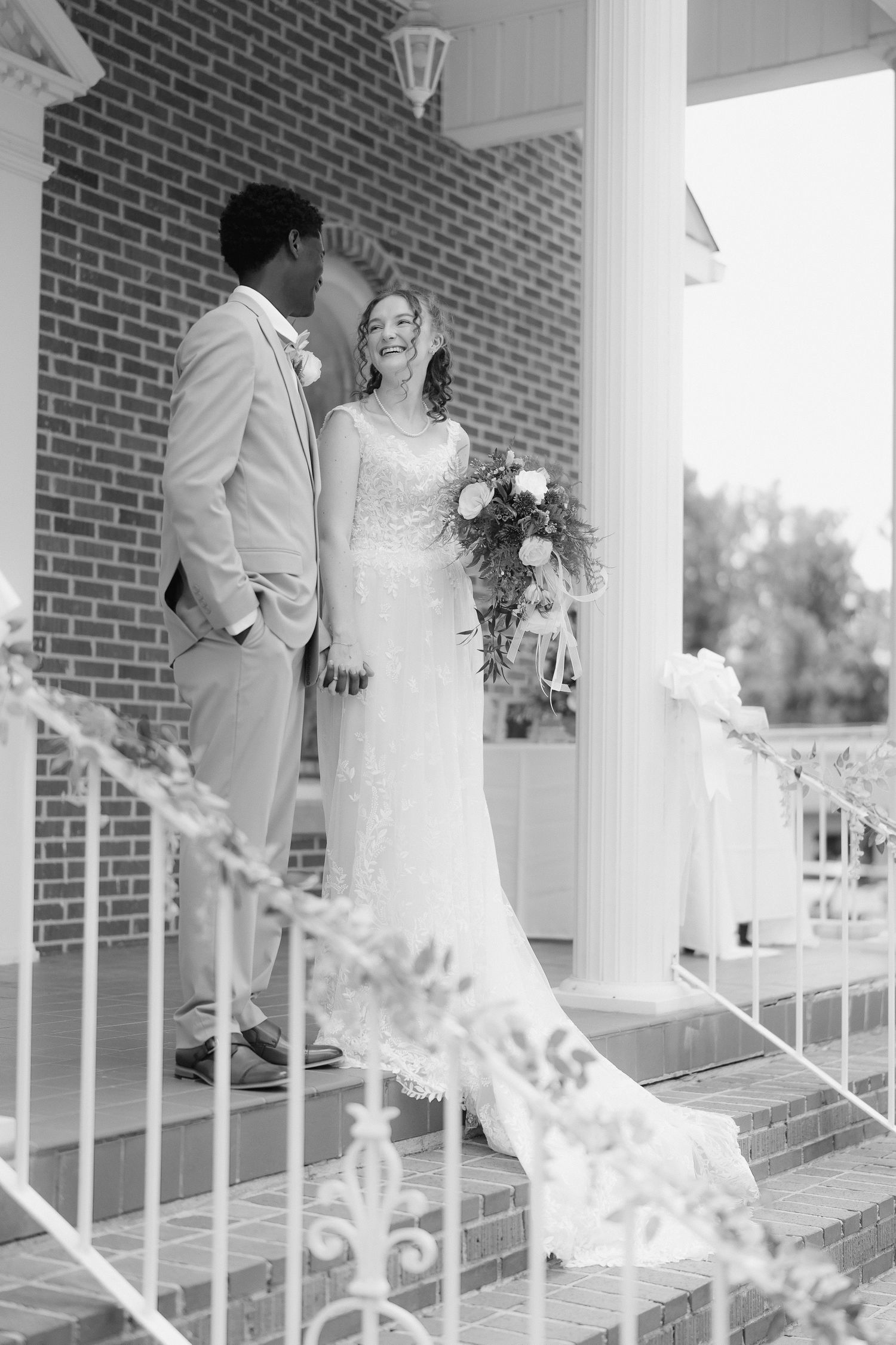 Black and white photo of smiling bride holding bouquet with groom on brick church steps.