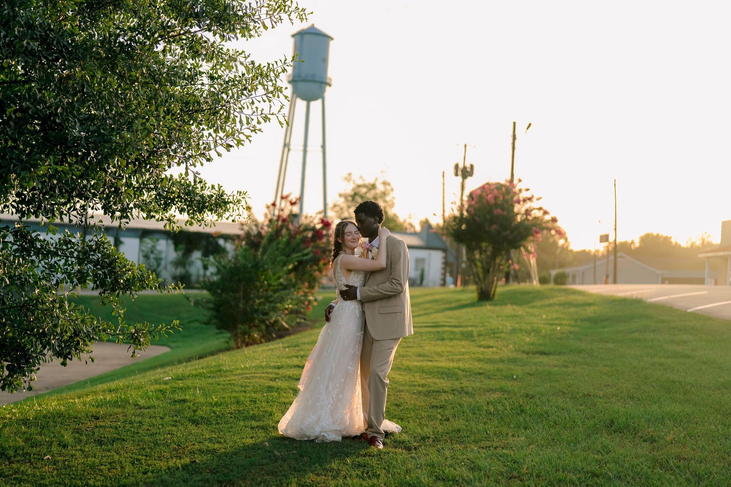 Newlywed couple sharing a romantic embrace on a green lawn at golden hour with a water tower backdrop.