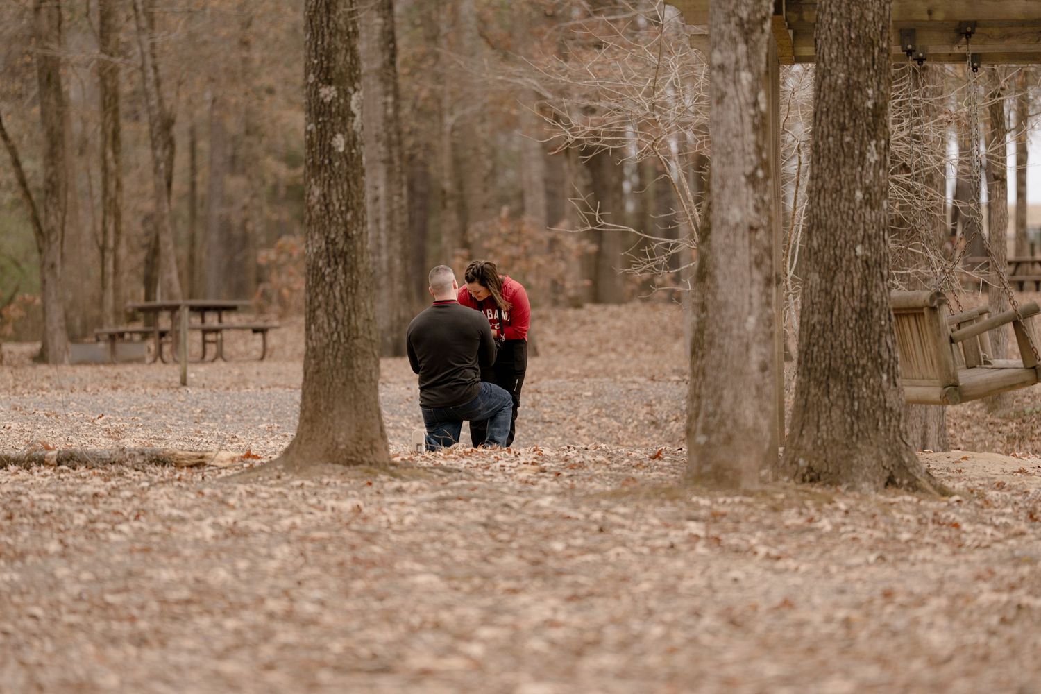 A person stands in a wooded area with bare trees and fallen leaves covering the forest floor in autumn.