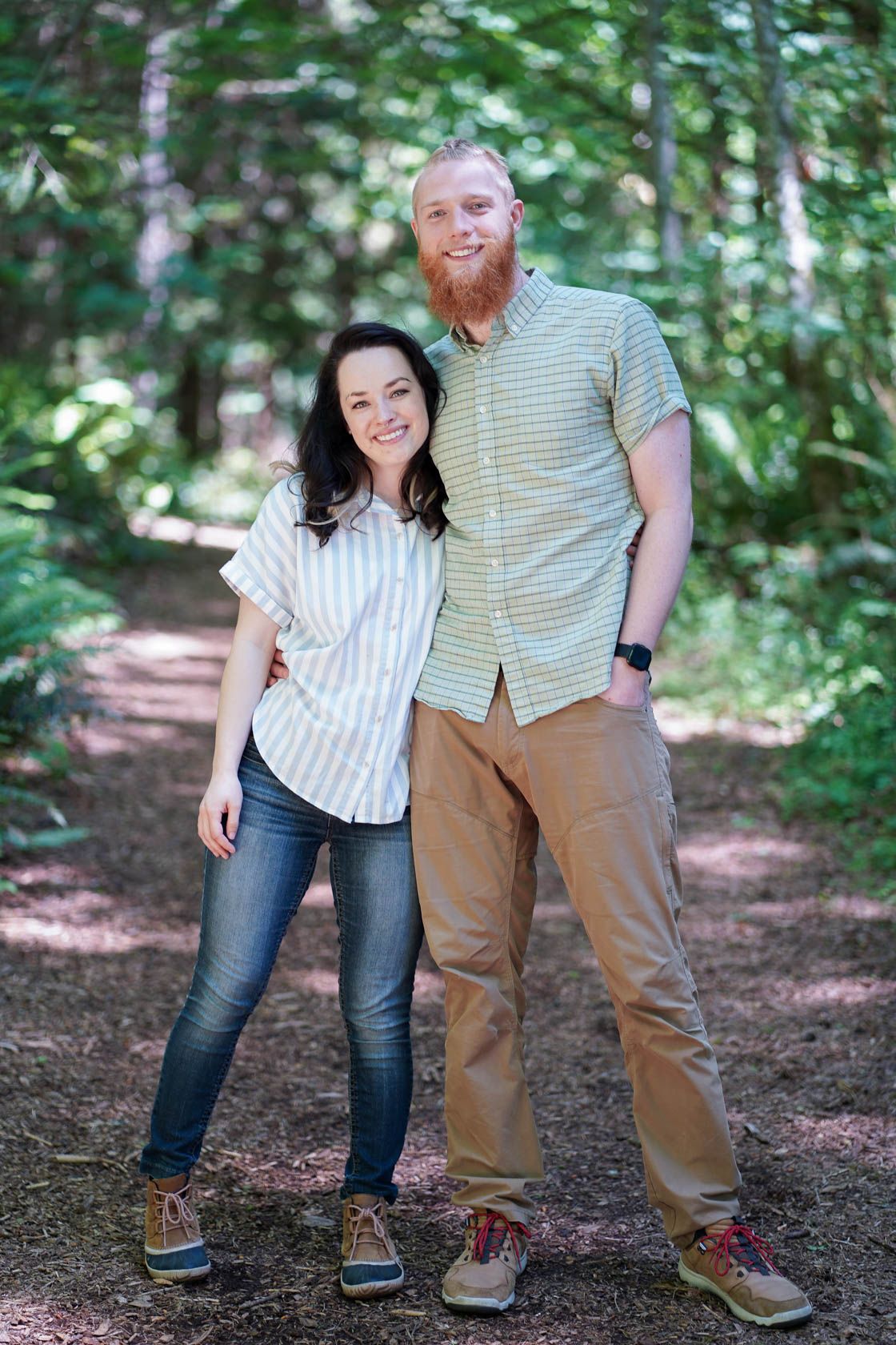 A casual portrait of two people in striped shirts standing on a wooded trail.