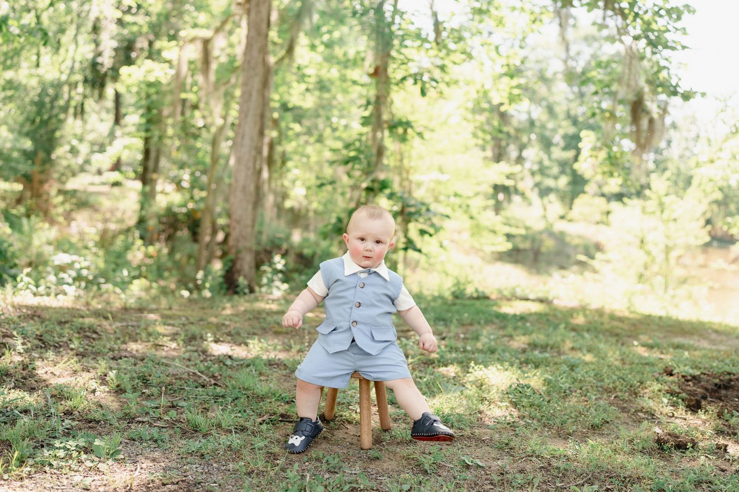 Small child in blue outfit sitting on wooden stool outdoors in a forest setting.