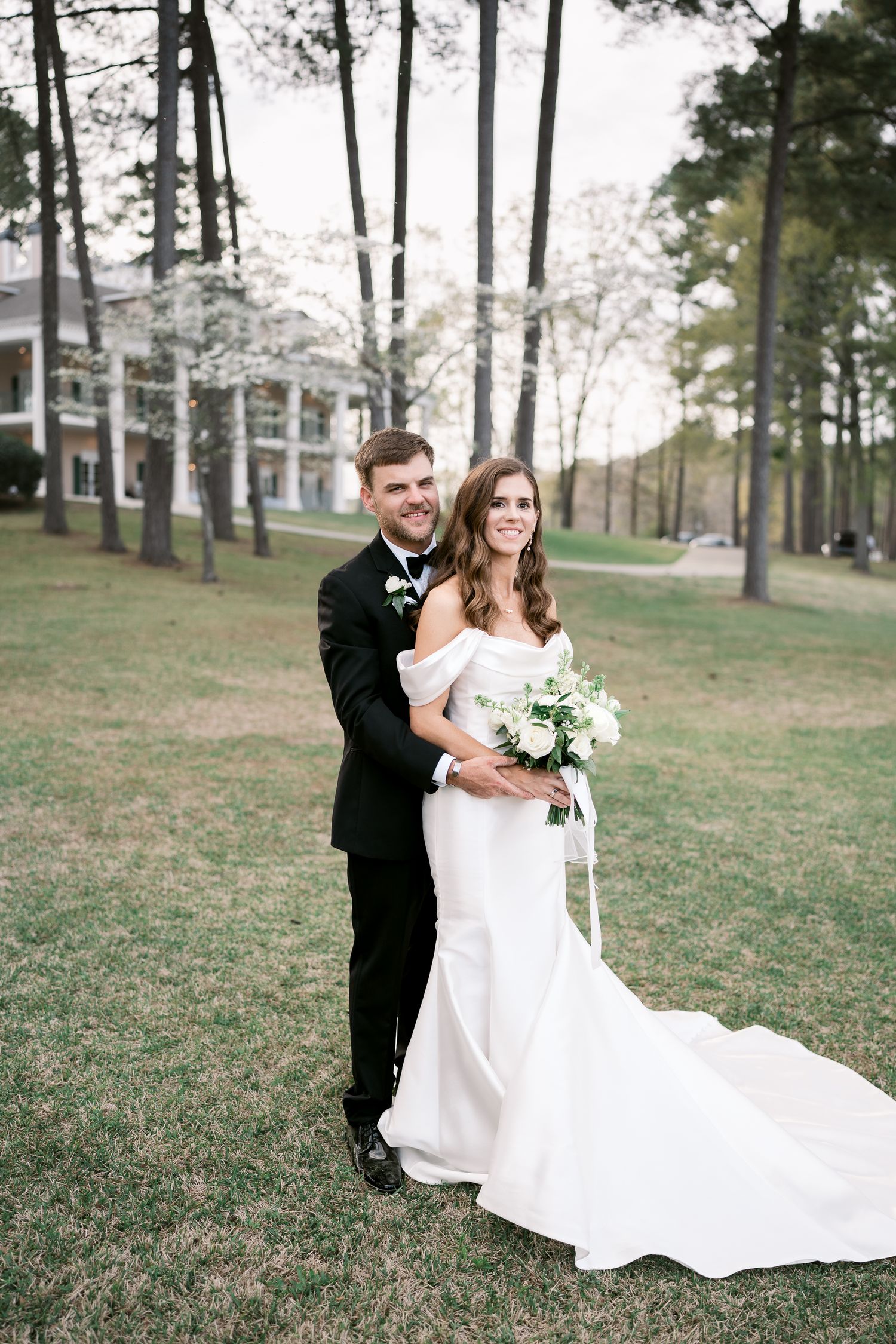 Newlyweds pose together on hillside with stately mansion and pine trees in background.