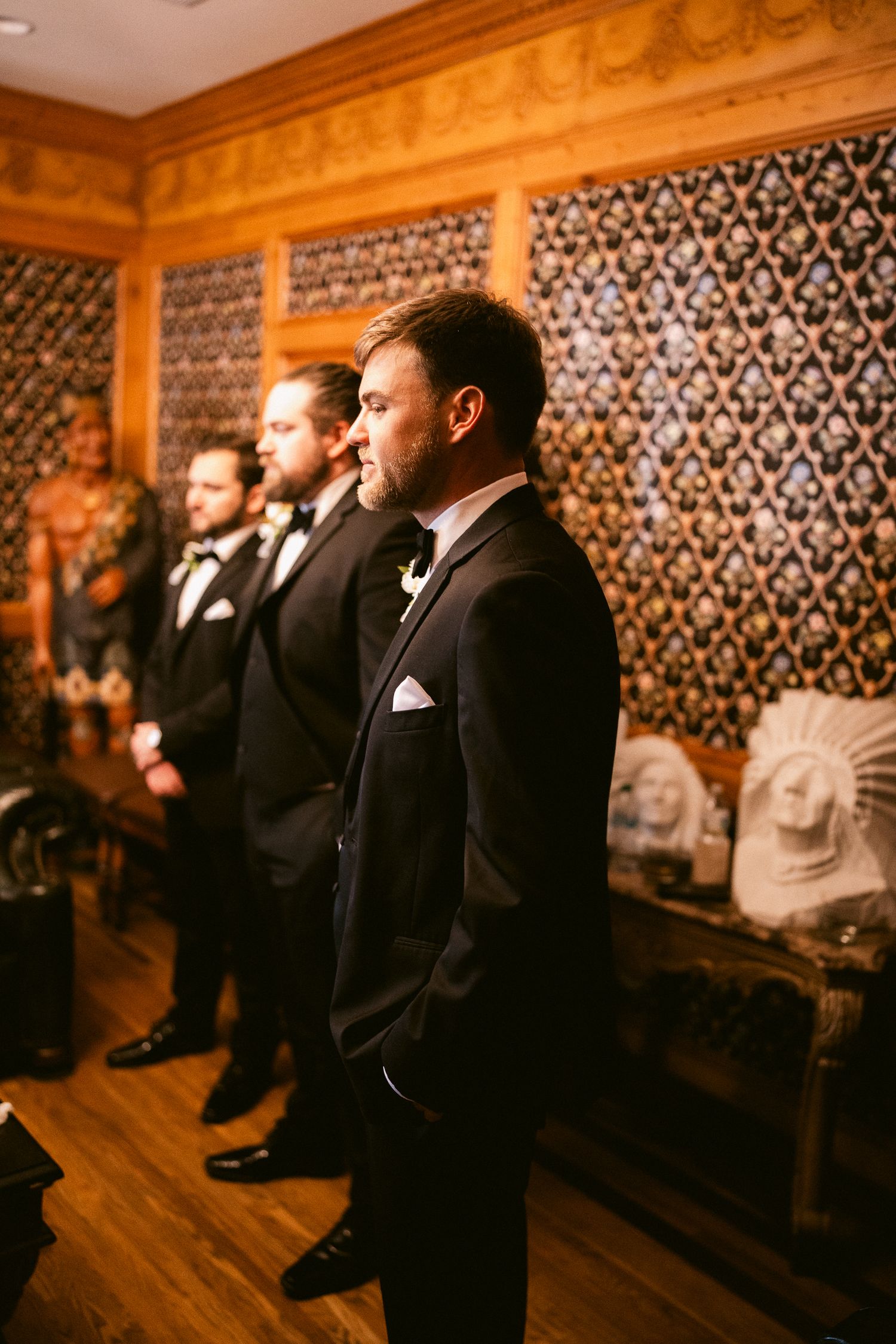 Groomsmen in black tuxedos stand in line against an ornate patterned wall during wedding ceremony.